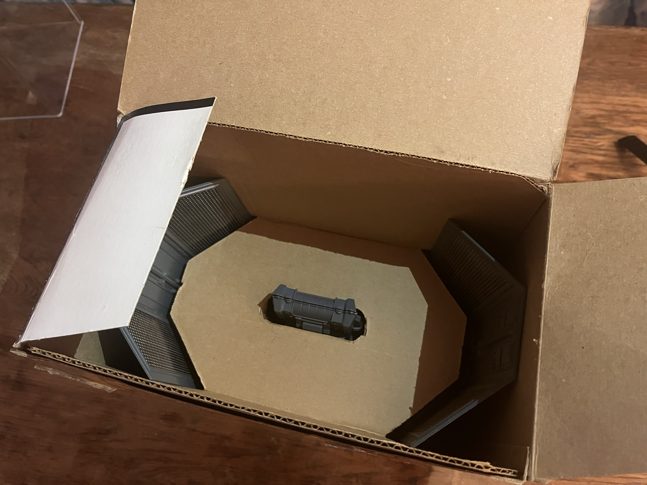 Open cardboard box with some black electronic devices and a piece of tan foam inside.