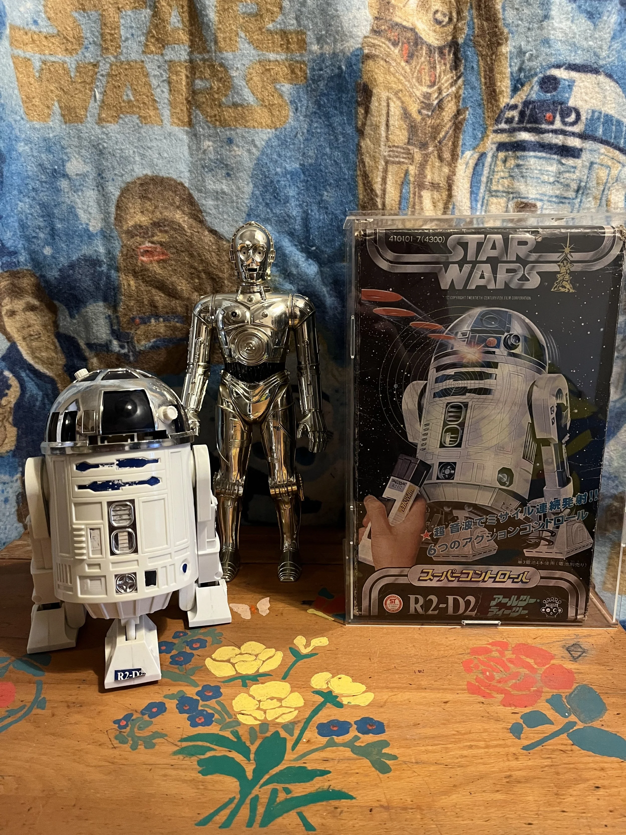 A collection of Star Wars memorabilia, including R2-D2 and C-3PO toys, with a Star Wars poster in the background, on a wooden table decorated with colorful floral patterns.