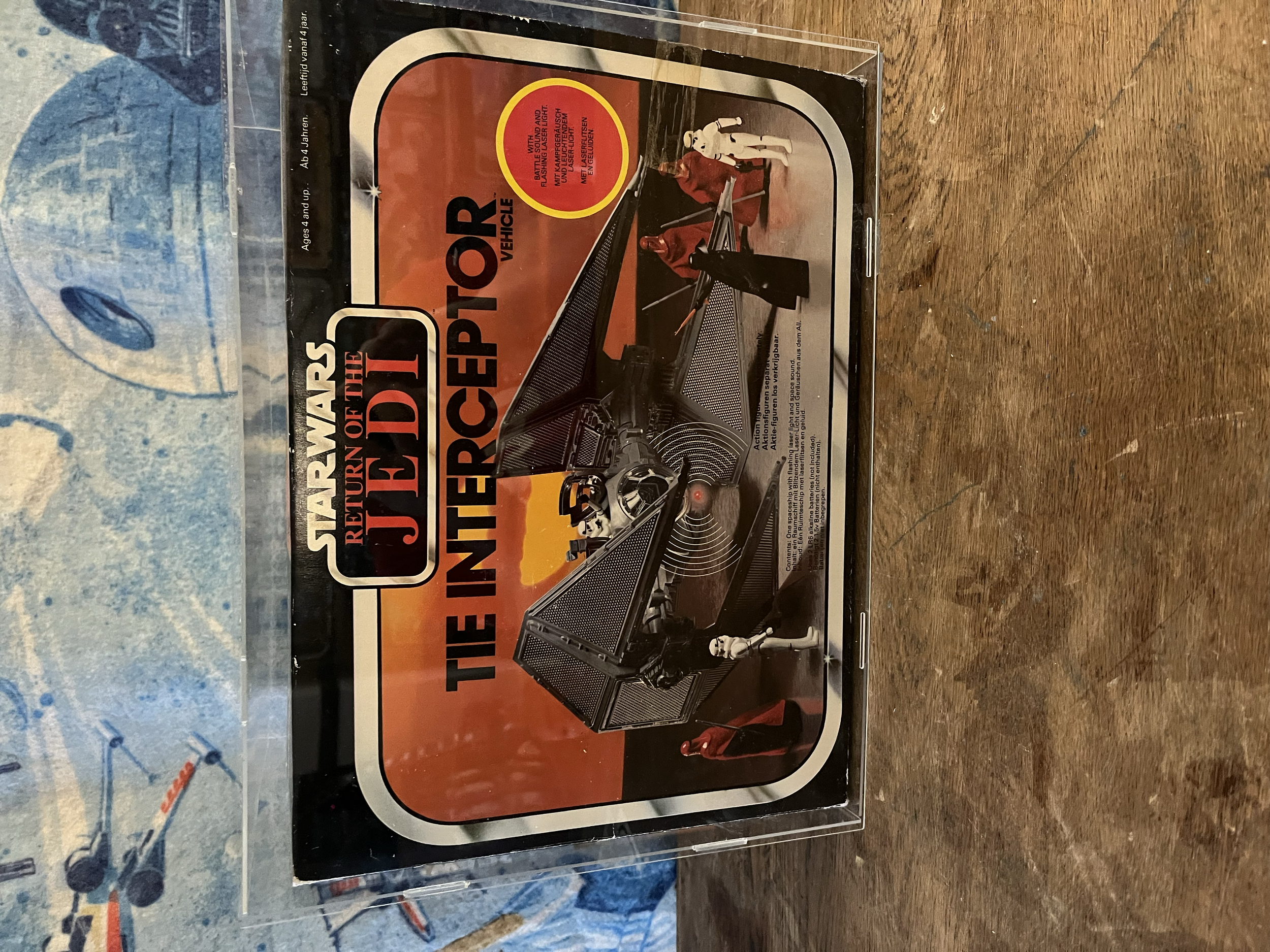 A toy set of a car with a space-themed design, featuring a white astronaut figure, is in its original packaging resting on a wooden surface with a blue, patterned background.