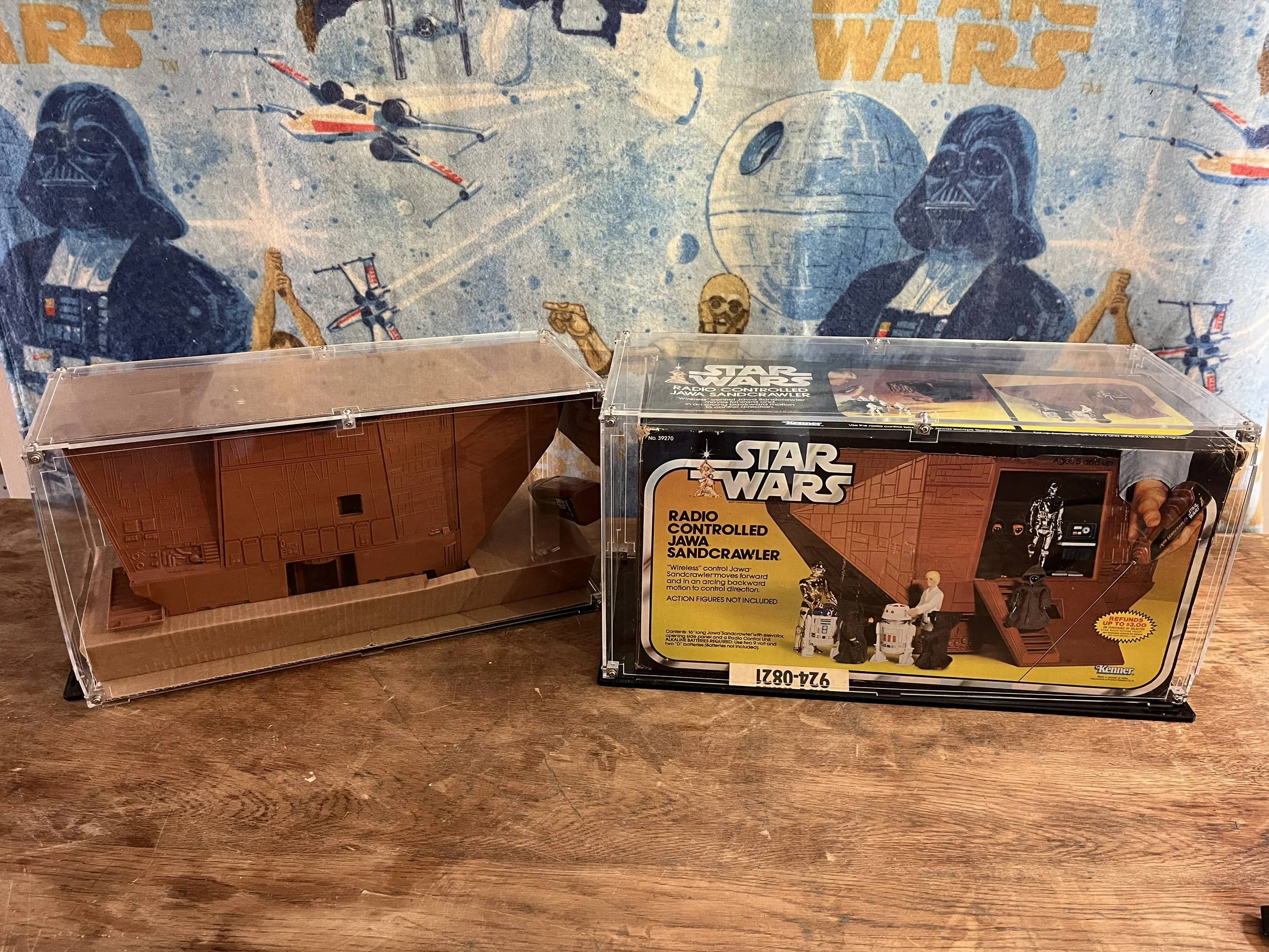 Star Wars Radio Controlled Jawa Sandcrawler toy set in a clear display case, with a vintage-style box showing images of the toy and action figures, with a large Star Wars themed wallpaper in the background.