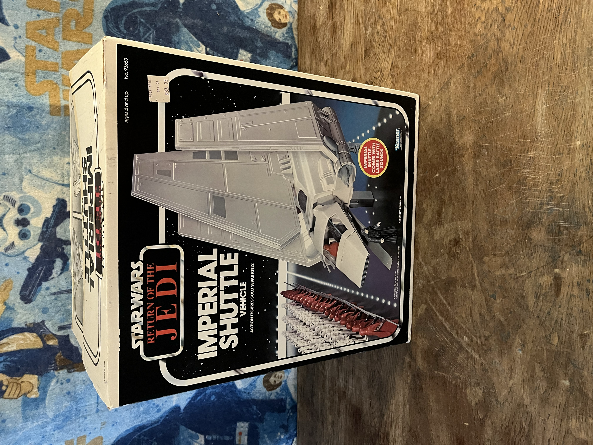 A toy model kit of a spaceship with instructions and packaging on a wooden surface, a blue patterned tablecloth partially visible in the background.