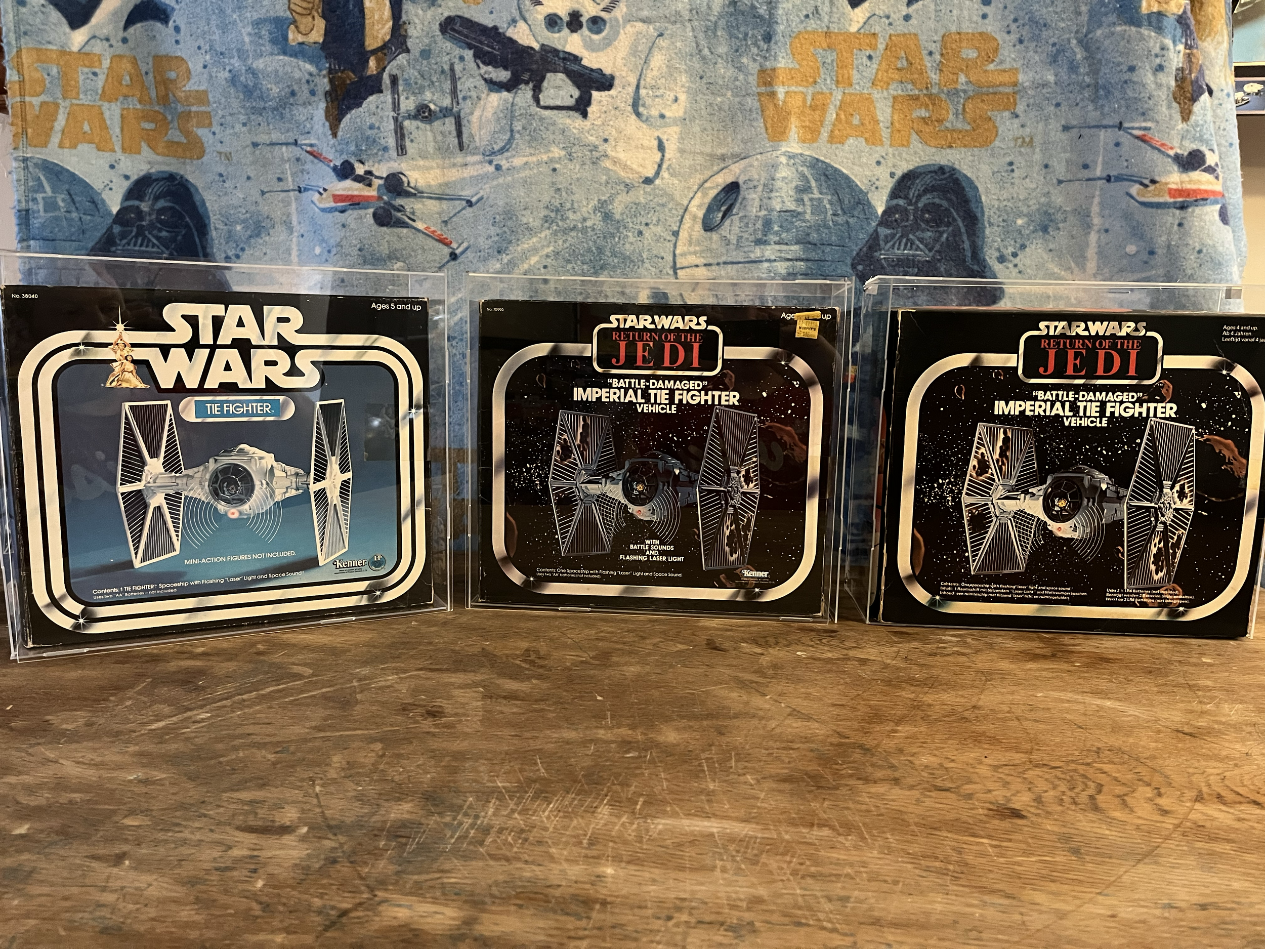 Three vintage Star Wars toy vehicle boxes displayed on a wooden surface, with a Star Wars themed backdrop featuring spaceships and characters from the franchise.
