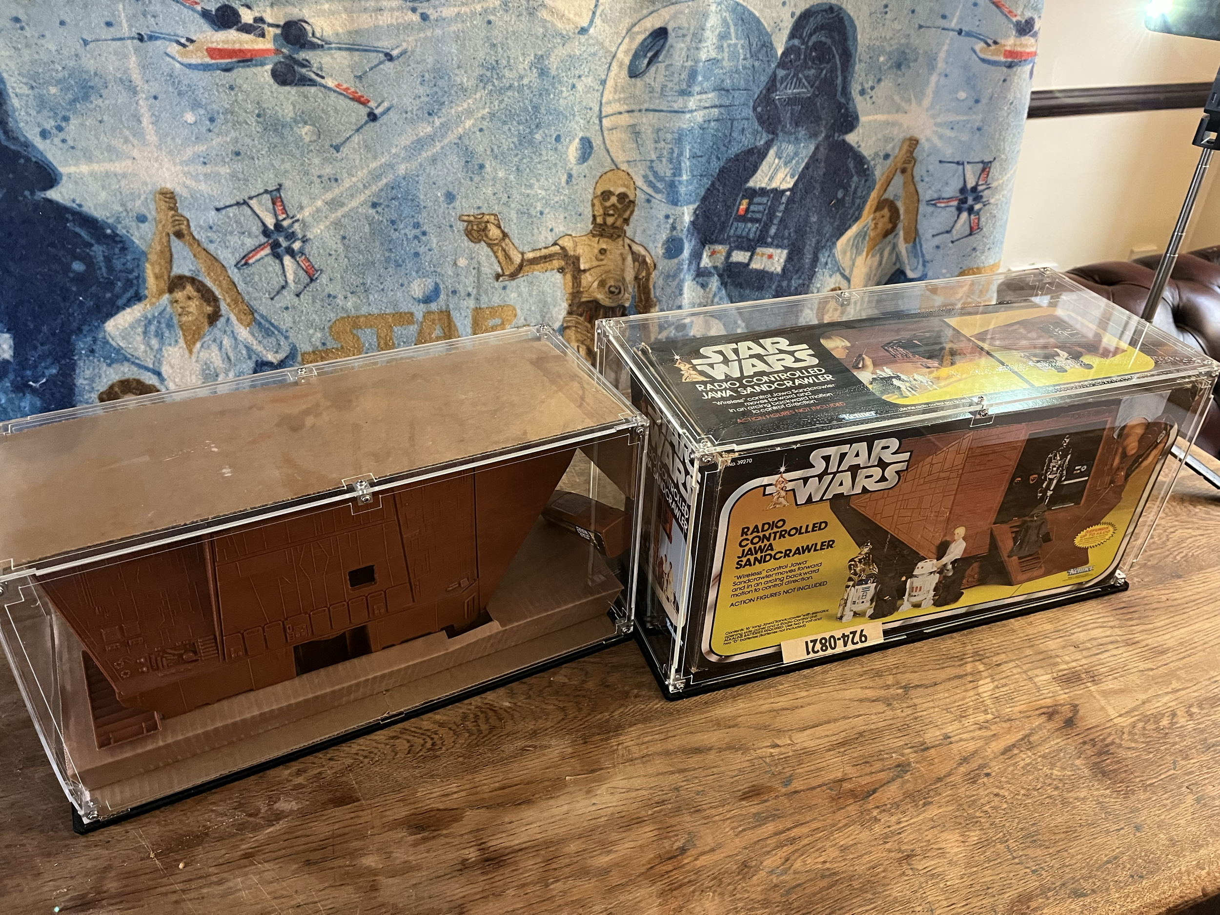 Star Wars action figure set in clear display case on a wooden surface with a Star Wars-themed fabric backdrop featuring characters and spacecraft from the series.