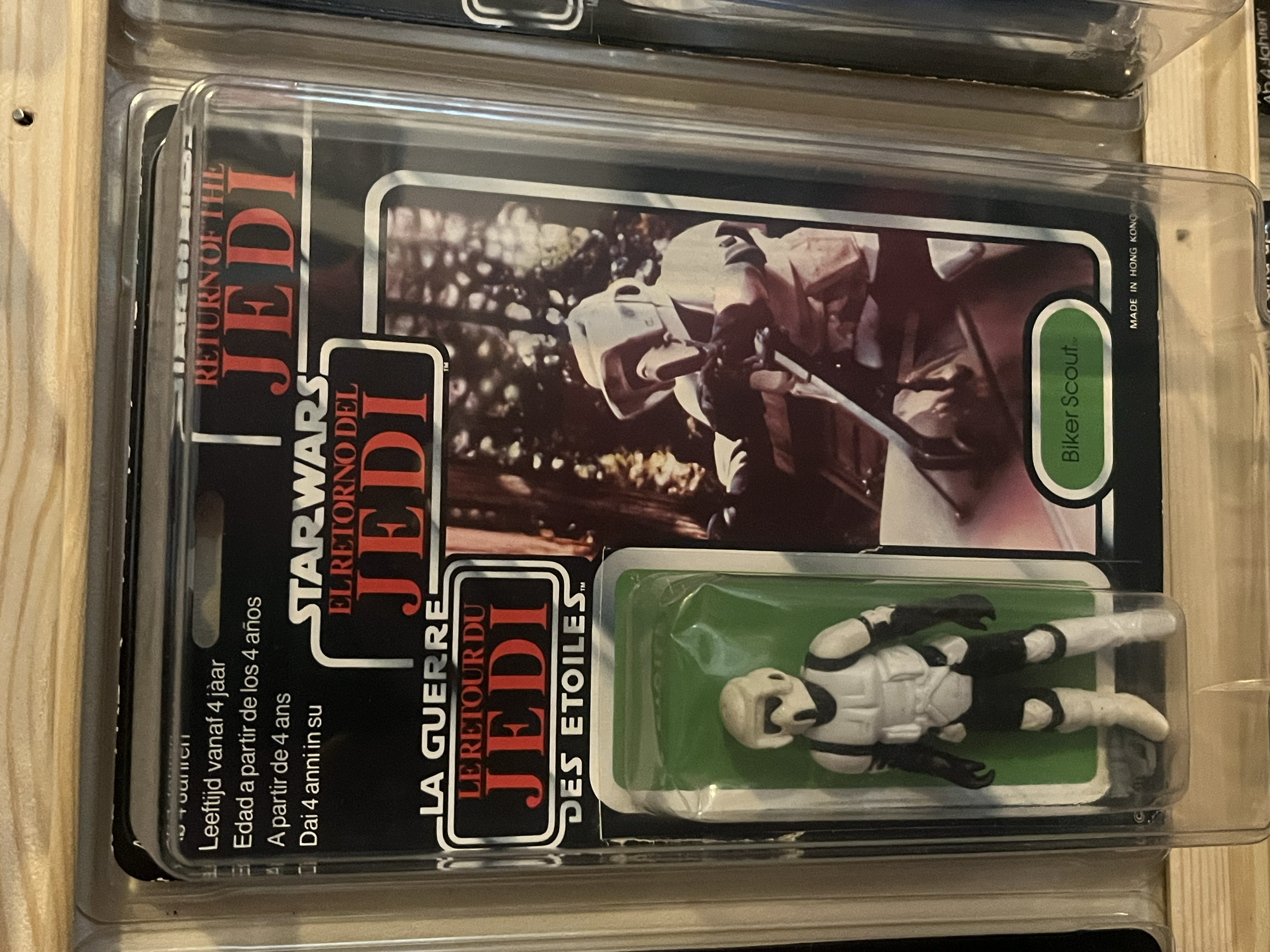 A clear plastic container holds a green toy scanner, a white toy skeleton, and a ghost mask. The container is placed on a wooden surface, and appears to be part of a display or storage for toys.