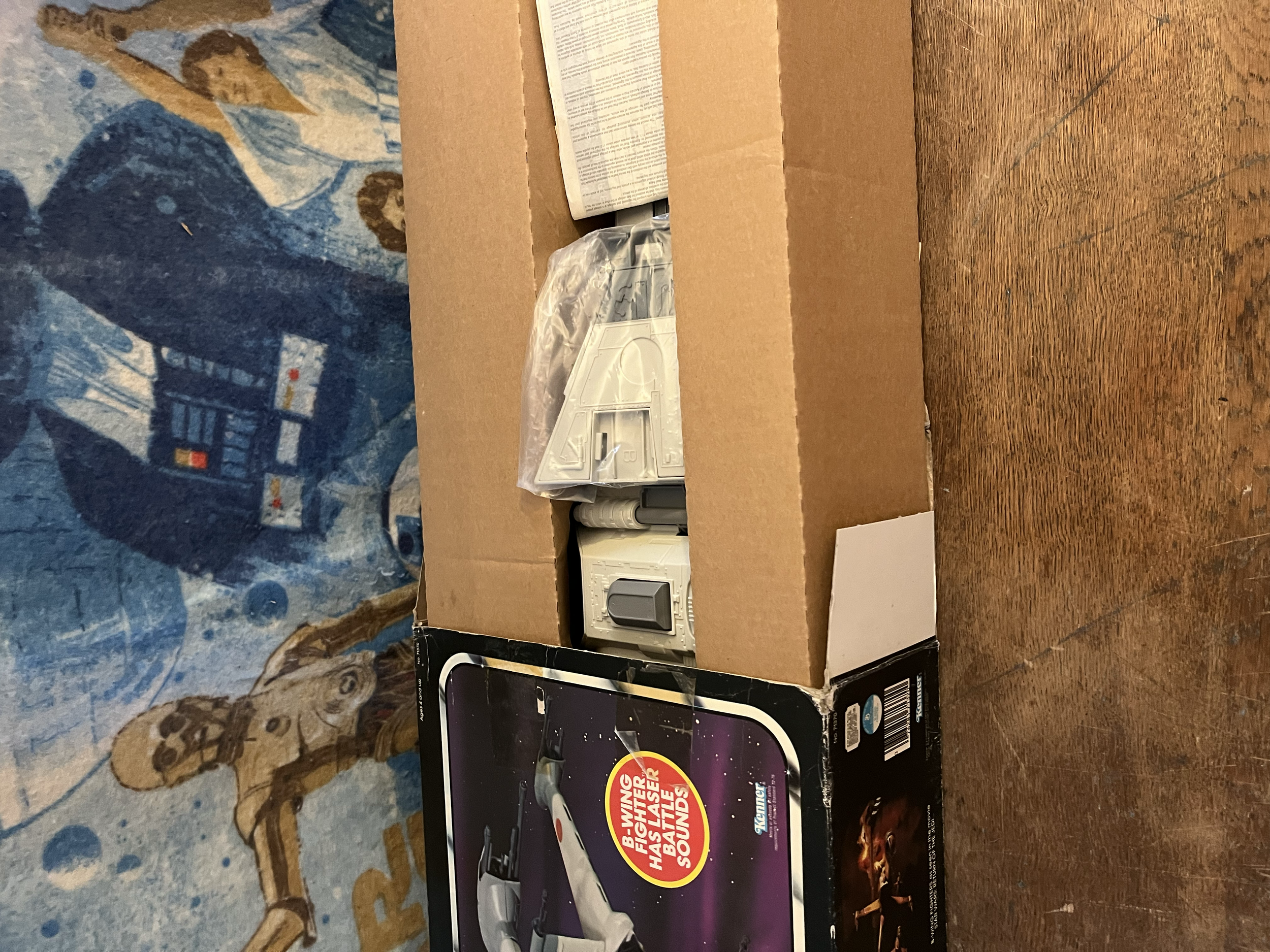A cardboard box containing various items including a video game case, plastic bag, and packaging materials, placed on a wooden surface with a blue patterned fabric in the background.