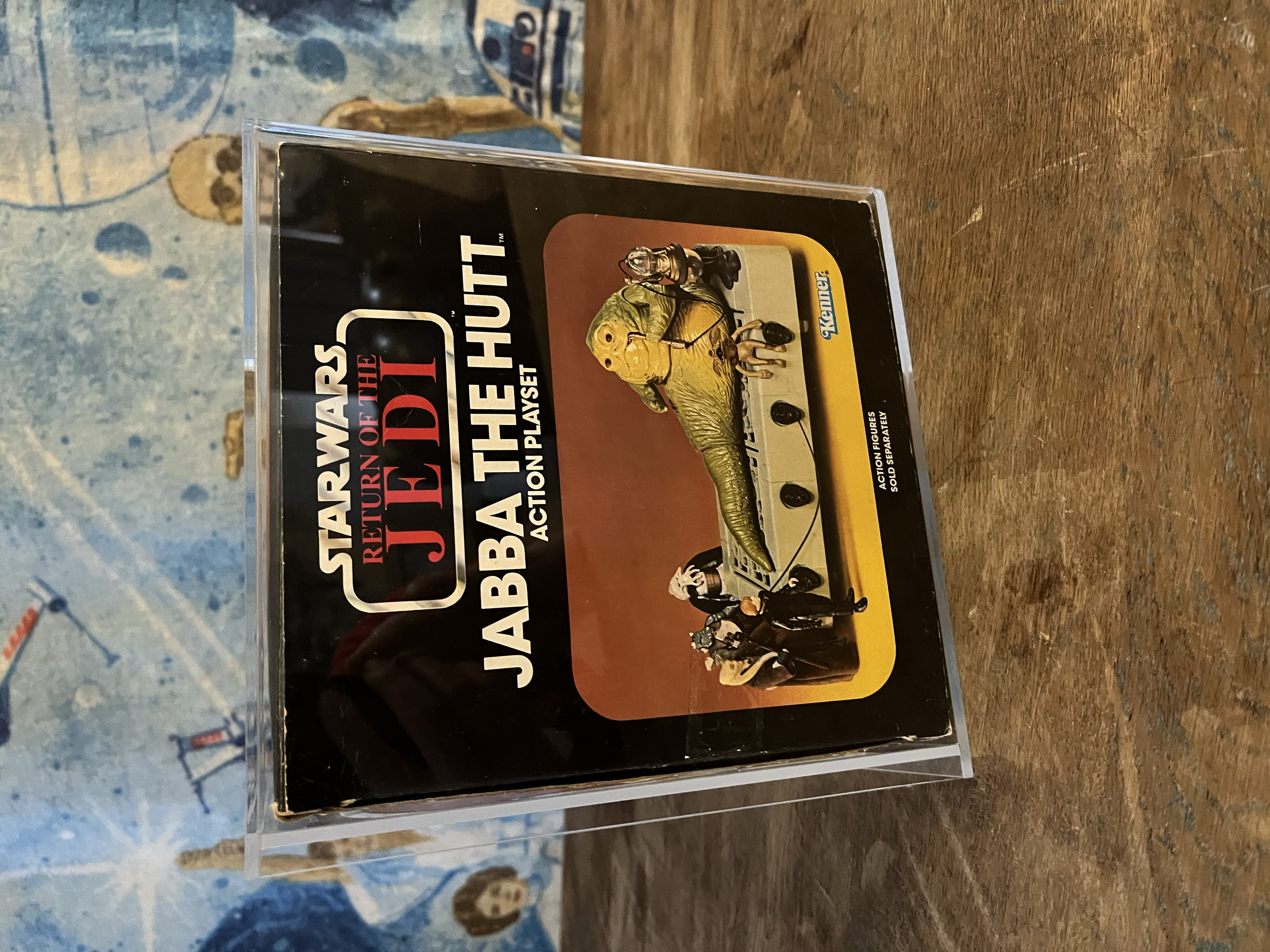 A toy set of the Jabba the Hutt character from Star Wars, packaged in a plastic case with a cardboard backing, on a wooden surface.