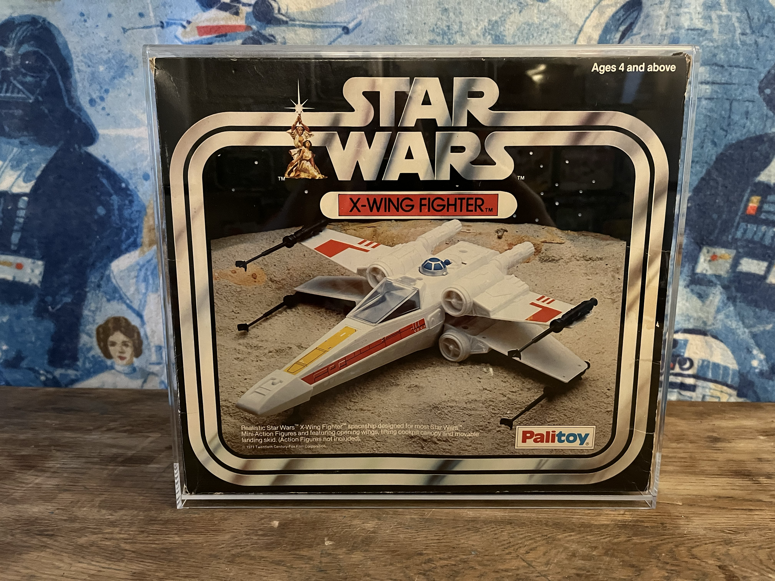 A miniature model of a Star Wars X-wing fighter spaceship inside a clear protective display case with a space-themed background and the Star Wars logo on the front. The case is placed on a wooden surface.