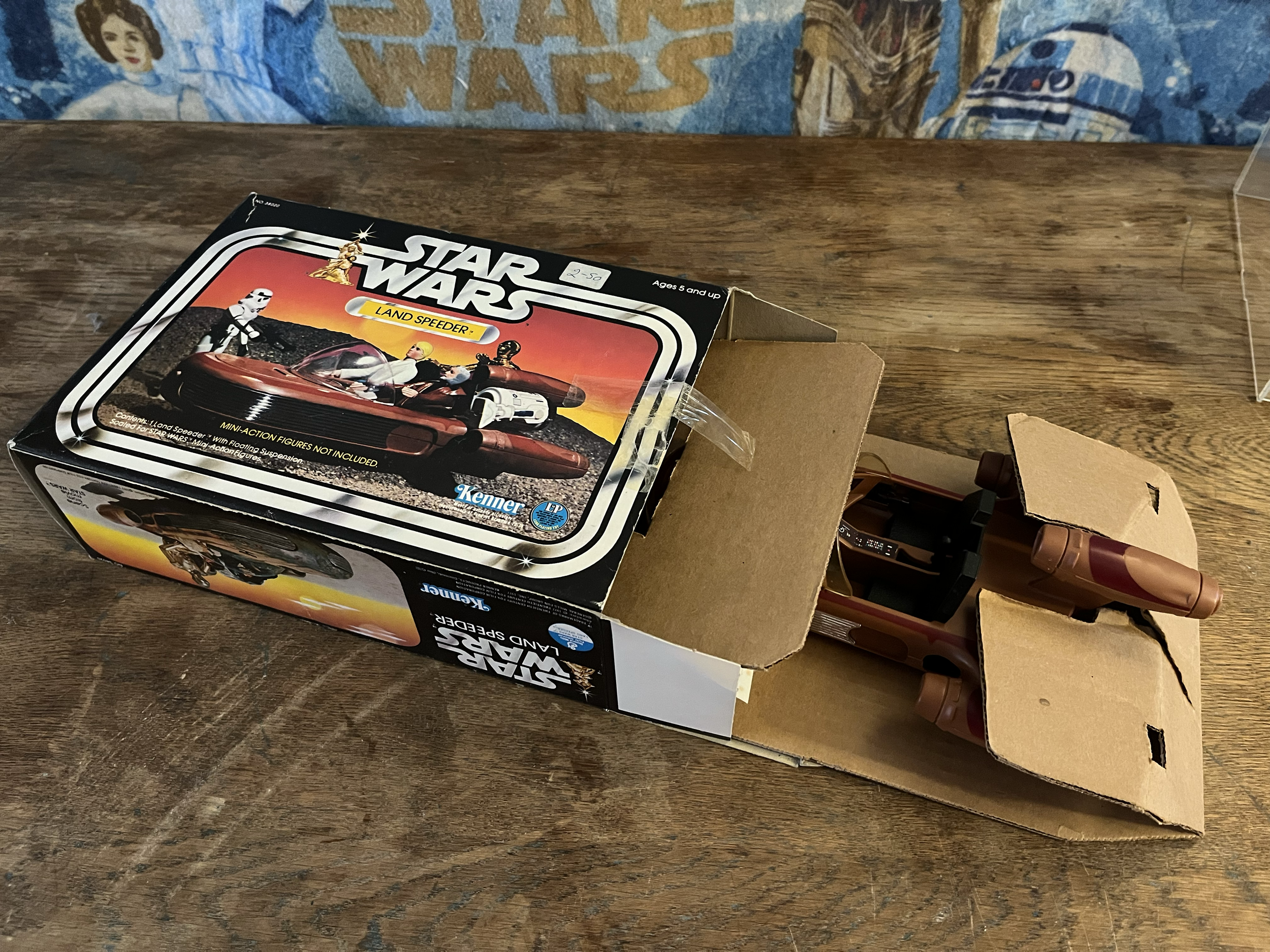 A vintage Star Wars toy set in a box labeled 'Land Speeder' on a wooden surface, with partial view of a Star Wars poster or fabric in the background.