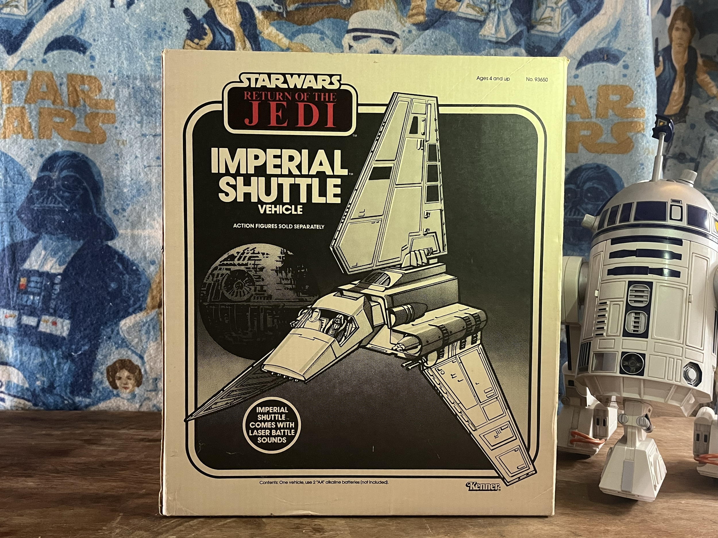 Star Wars Return of the Jedi Imperial Shuttle vehicle toy box with illustration of the shuttle and R2-D2 toy figure beside it. Background includes a Star Wars-themed fabric and some model figures.