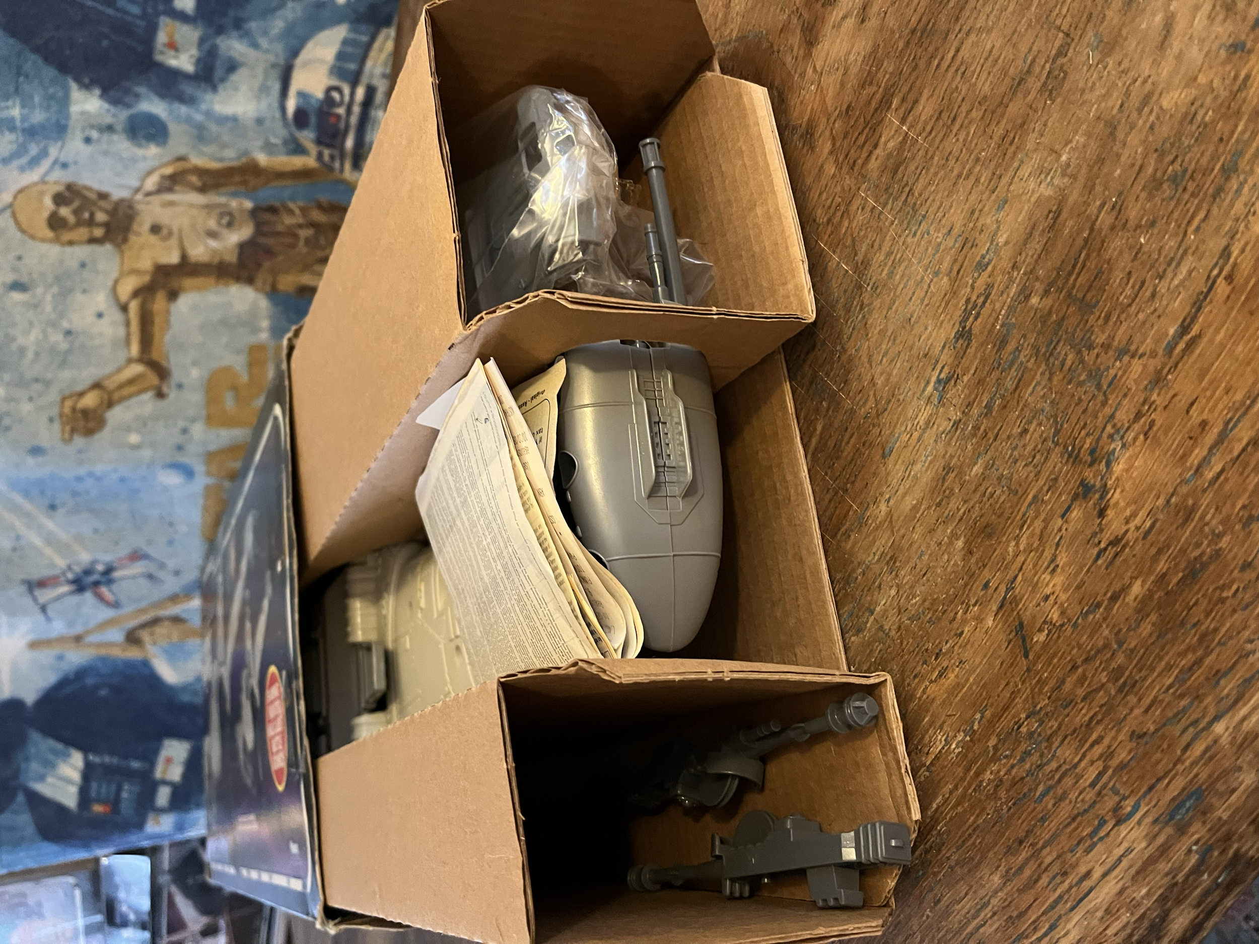 A cardboard box containing various gray plastic parts and manuals on a wooden surface.