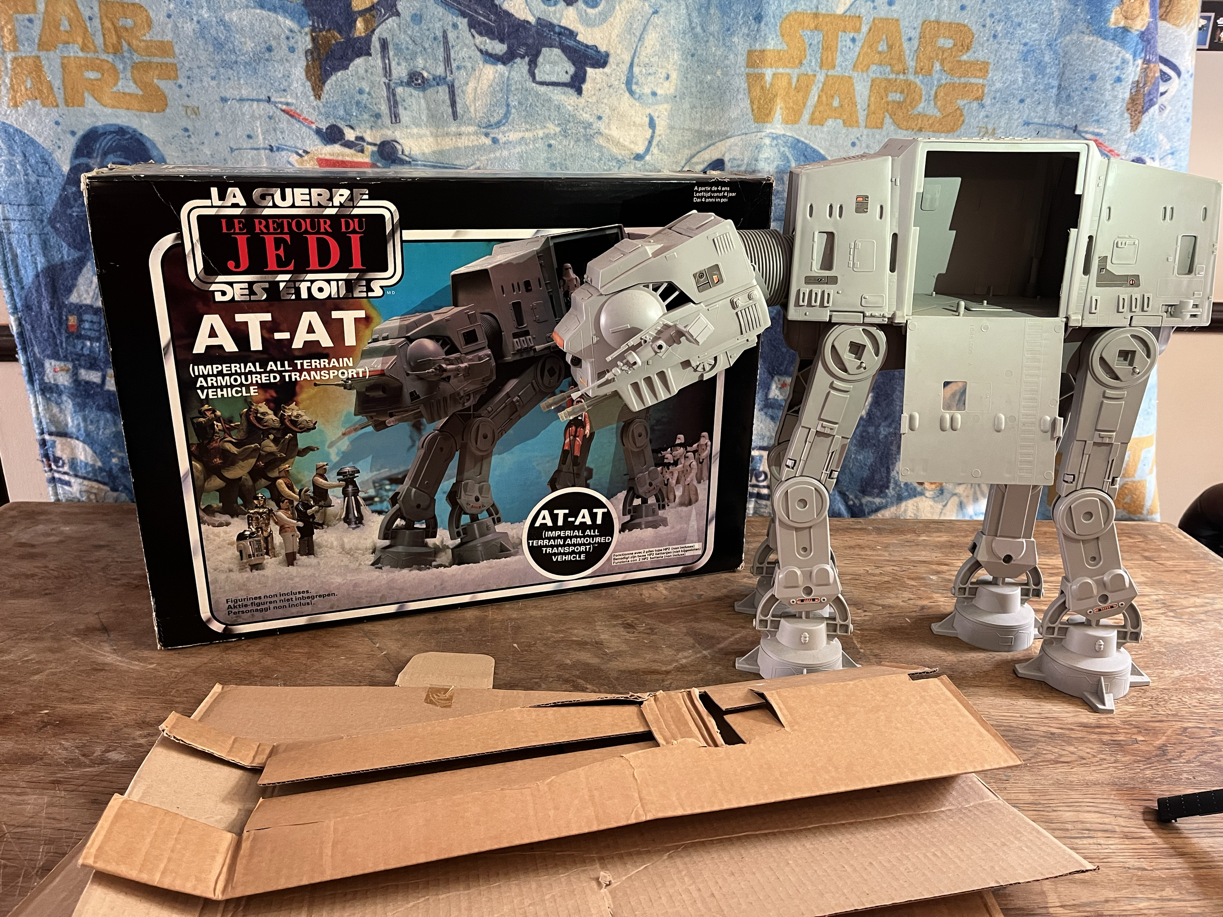 A vintage Star Wars toy set featuring the AT-AT (Imperial All Terrain Armoured Transport) vehicle, with the box showing an illustration of the toy and some miniature figures, was placed on a wooden table with an opened cardboard box in front of it. T