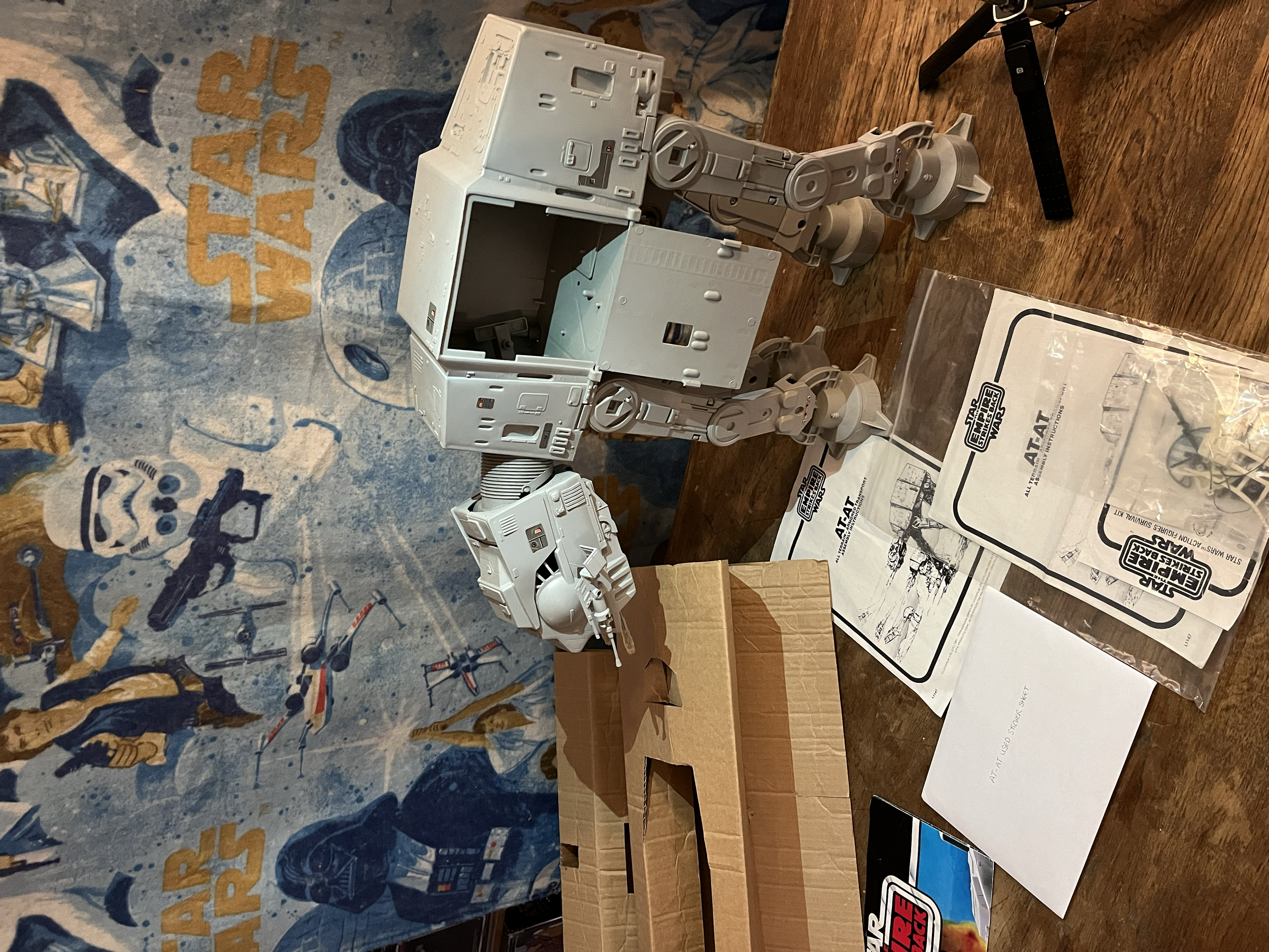 Parts of a Star Wars robot model assembly on a wooden table, with instruction manuals and tools nearby, against a Star Wars-themed carpet background.