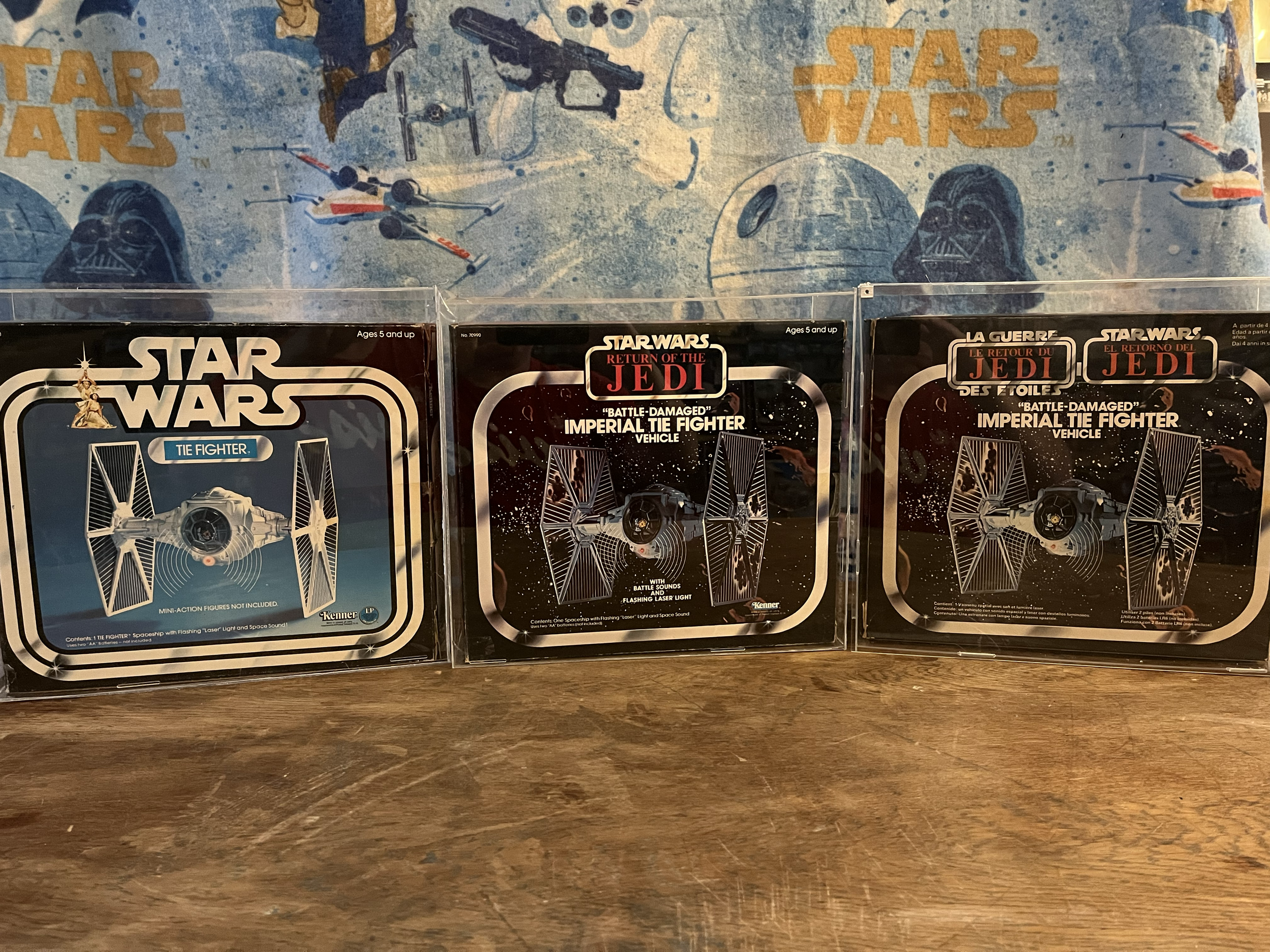 Three vintage Star Wars Kenner Tie Fighter packaging boxes featuring Imperial TIE Fighter vehicles displayed on a wooden surface. The background has a blue Star Wars-themed wallpaper with images of Darth Vader, TIE Fighters