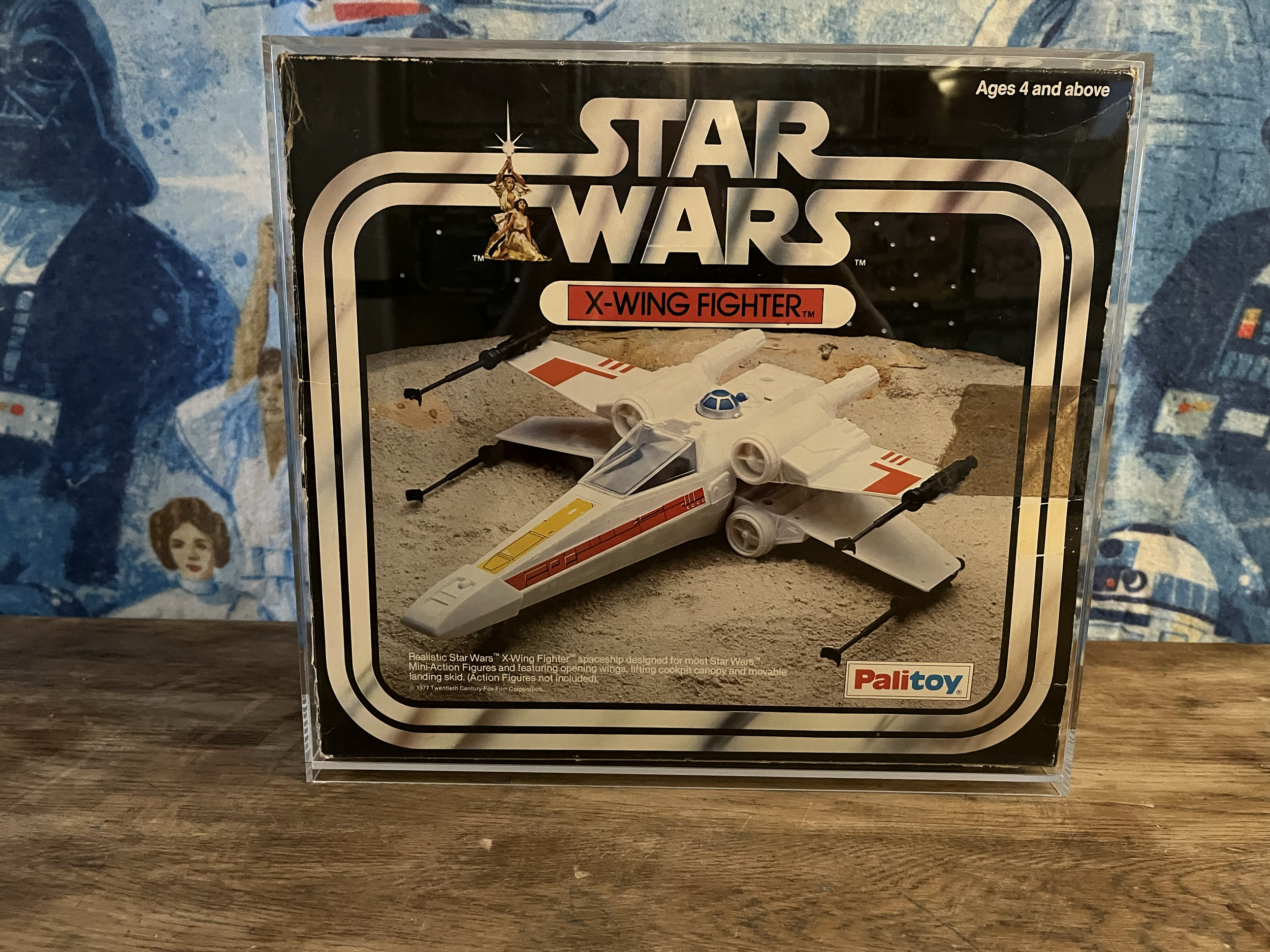 Star Wars X-Wing Fighter miniature in its packaging, featuring a spaceship designed for the Star Wars franchise, with a background showing a desert landscape and a starry sky.