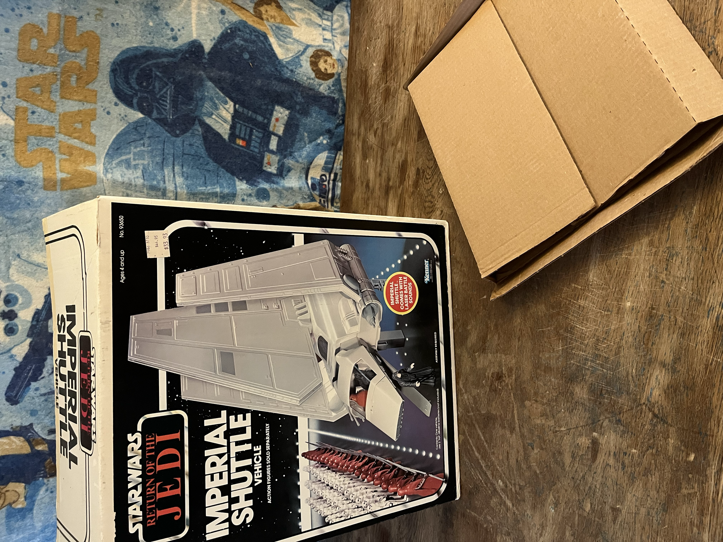 A boxed set of miniature military model figures and equipment, including a gray tank, red and white soldiers, and a display with guidance on assembly and painting, positioned on a wooden surface with a colorful background featuring a blue poster with