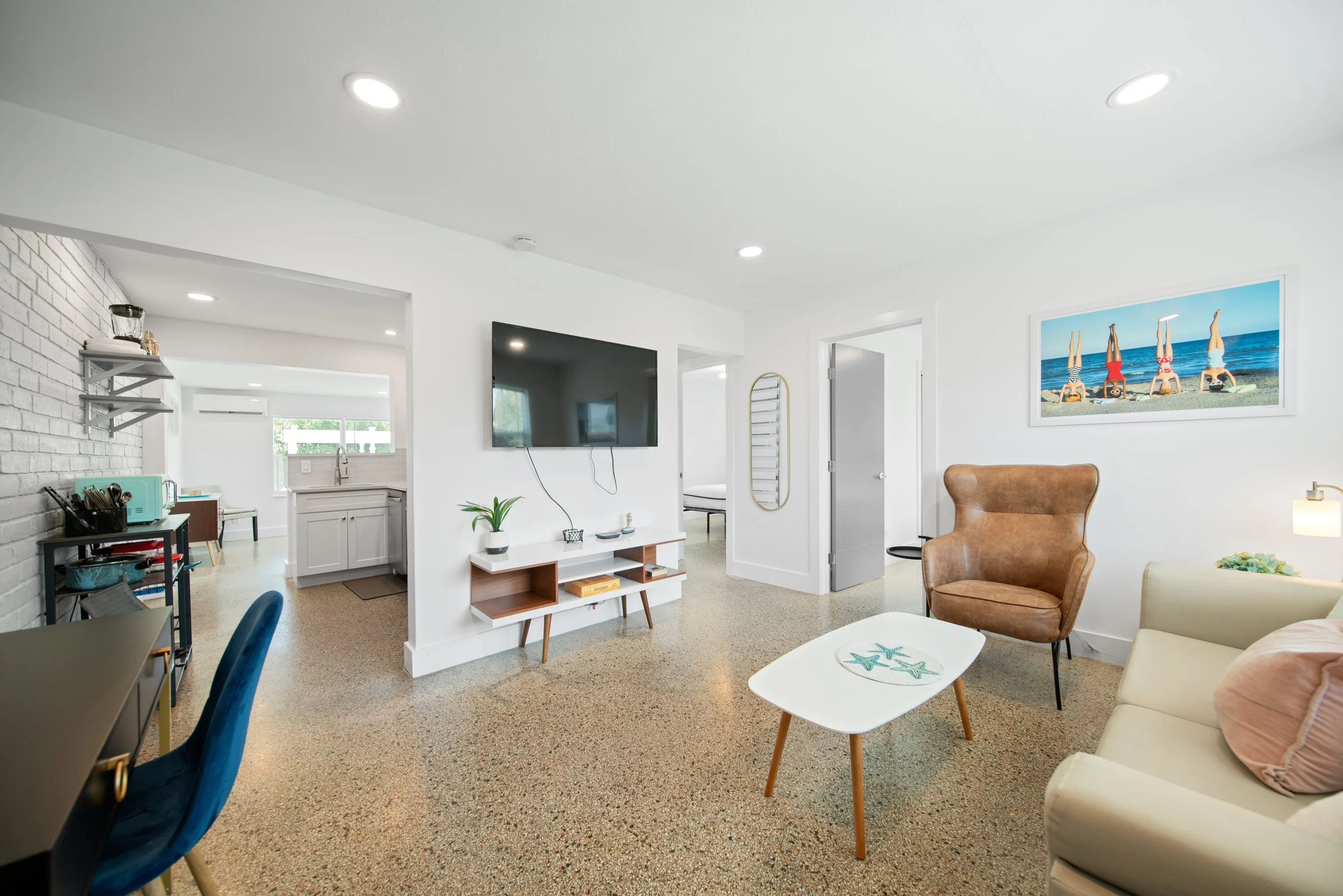 A modern living room with white walls and a ceiling with multiple recessed lights. There is a large flat-screen TV mounted on the wall, and below it, a white console table with decorative items. To the right, there's a large framed photograph of chil