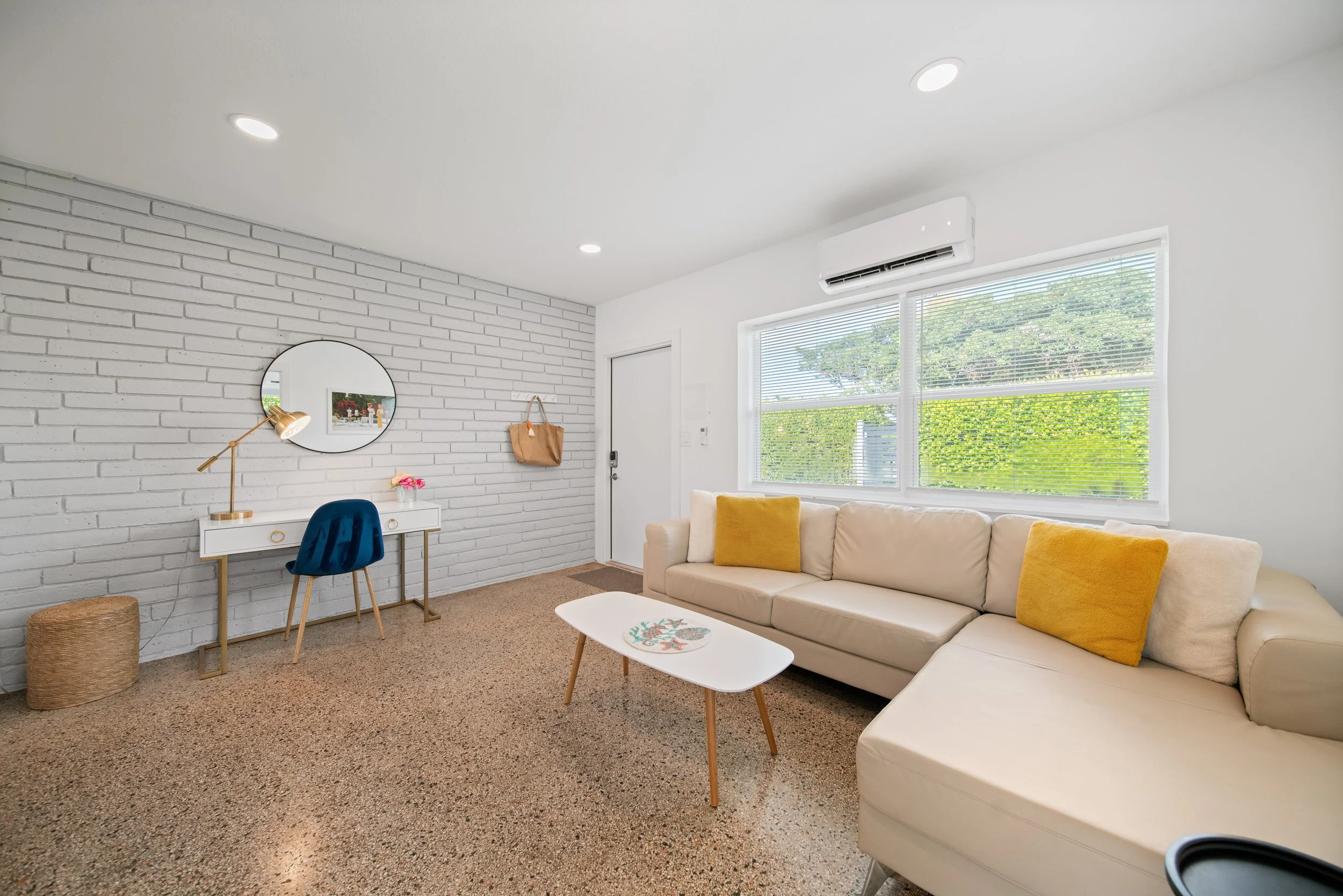 Bright living room with white brick wall, beige sofa with yellow pillows, small white coffee table, window with blinds, and ceiling air conditioner.