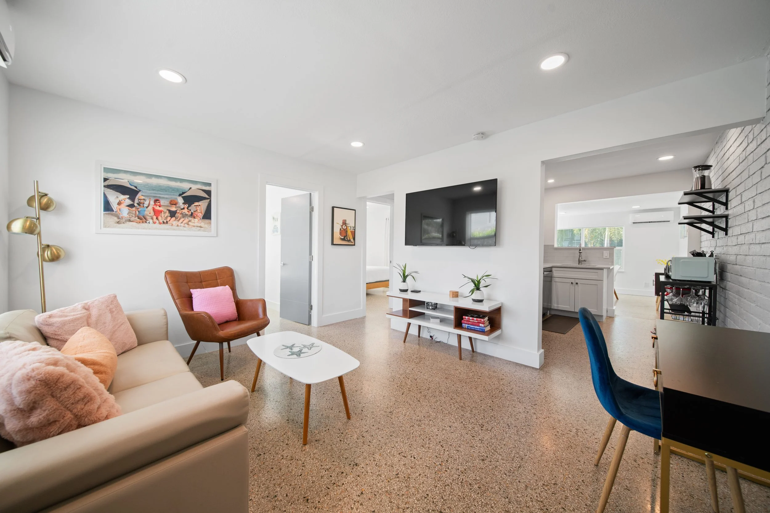 Bright living room with tan sofa, pink pillows, brown leather armchair, white coffee table, and wall art of children at the beach. Open view to kitchen with white cabinetry and grey brick wall with black shelves.