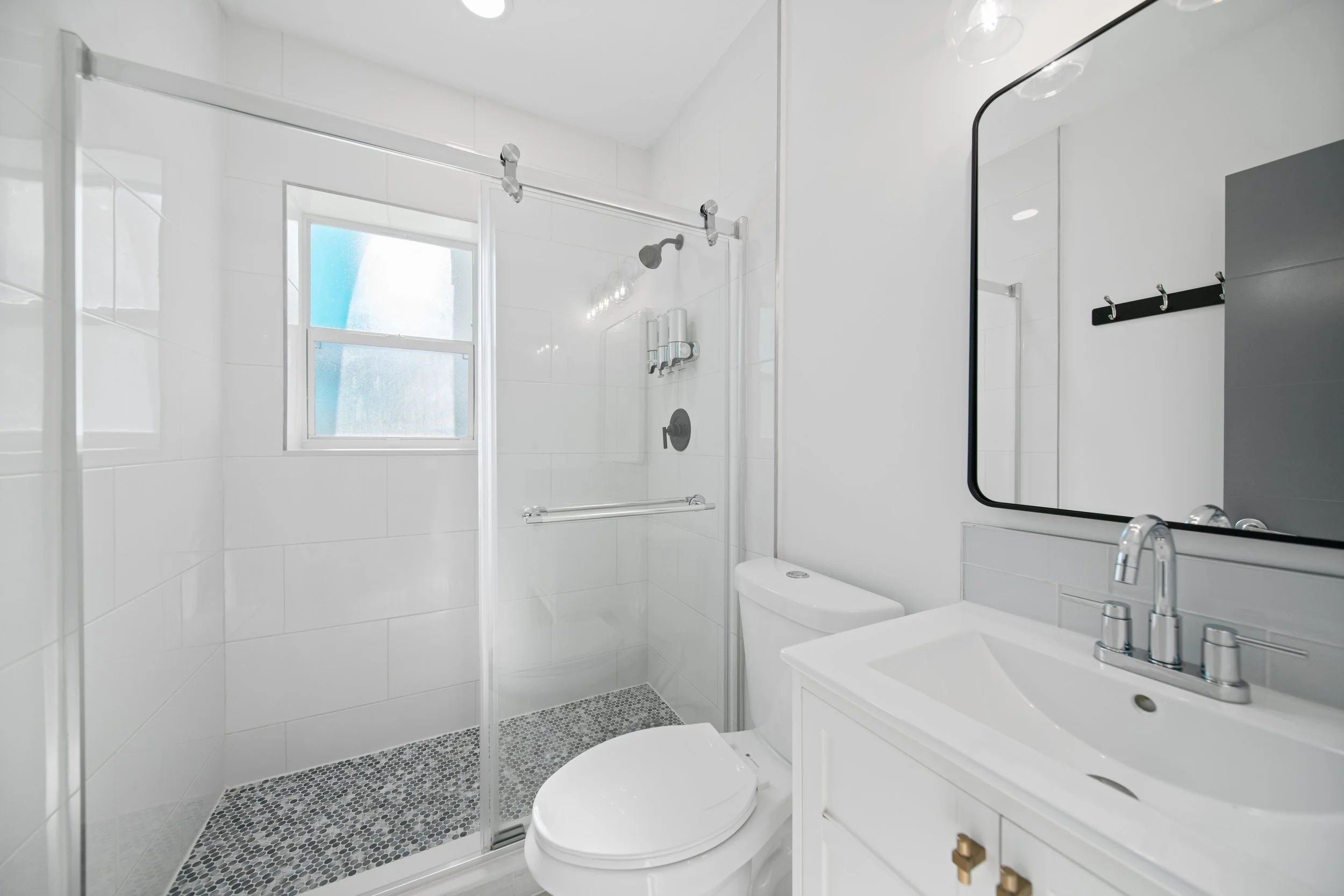 A modern, white-tiled bathroom featuring a walk-in shower with a glass door, a window, a wall-mounted showerhead, and a small pebble-textured floor in the shower area. There is a white toilet and a white vanity with a sink in the foreground, and a mi