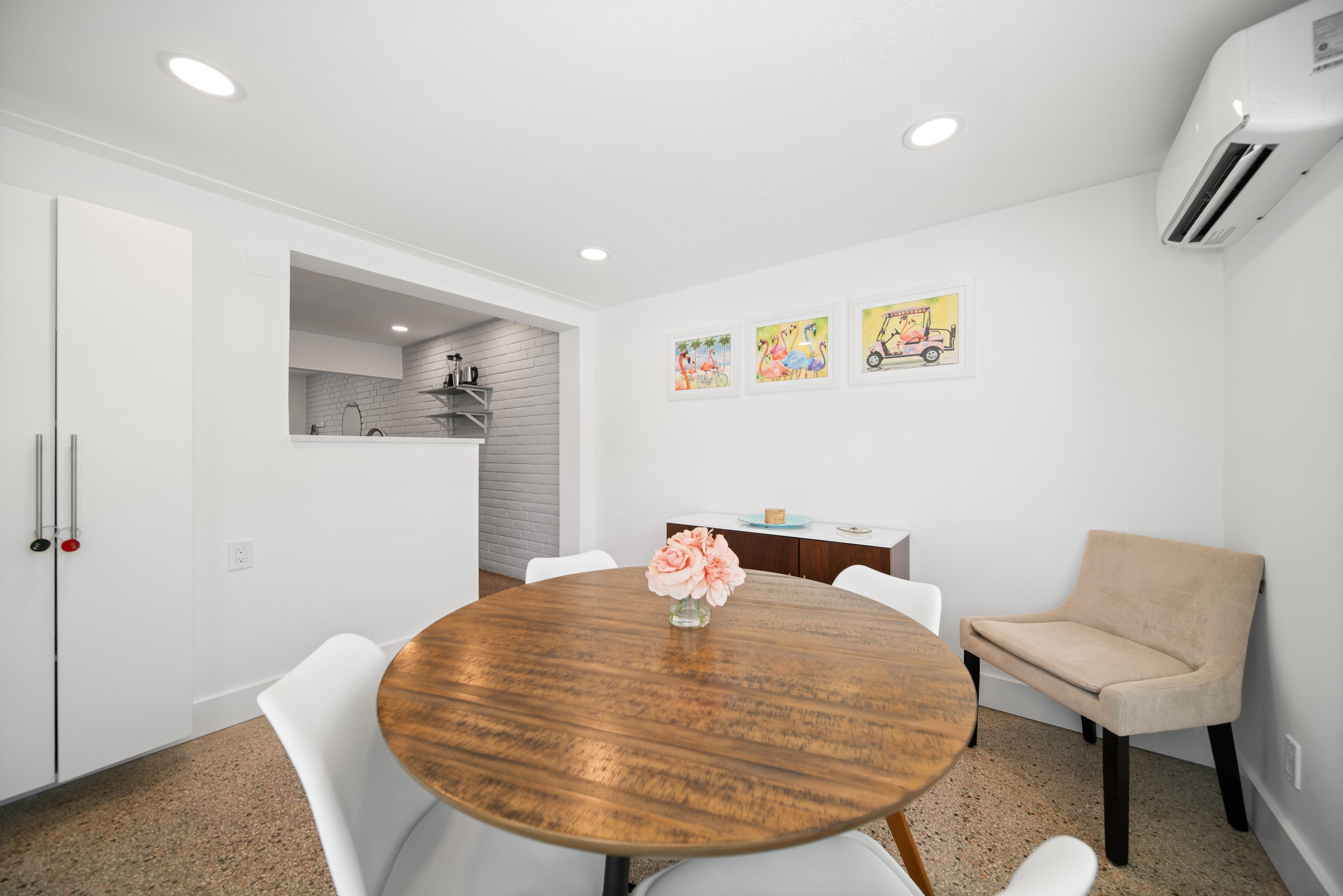 A dining area with a round wooden table, white chairs, and a beige cushioned bench. A vase with pink flowers is on the table. The wall behind has three colorful flamingo-themed pictures, and there is an air conditioning unit on the upper right wall.