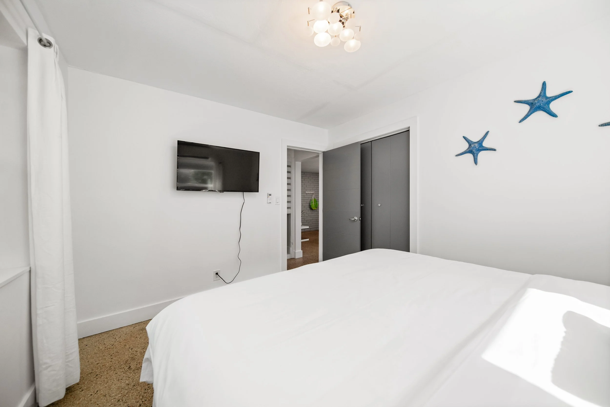 A bedroom with white walls, a bed with white bedding, a mounted flat-screen TV, a modern chandelier ceiling light, a doorway leading to a hallway, and blue starfish wall decorations.