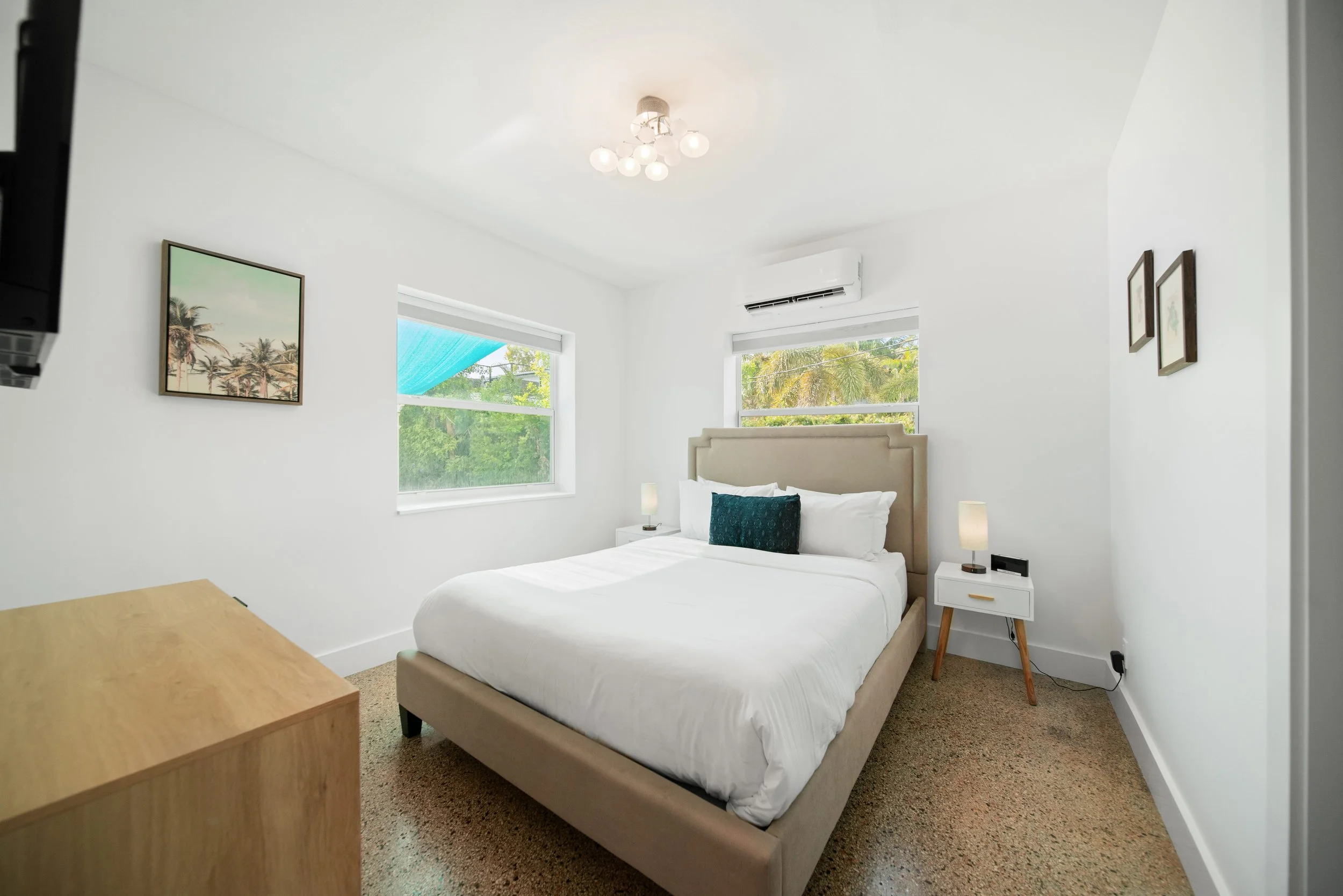 Bright bedroom with white walls, a beige bed with white bedding and dark green pillow, wooden nightstand with lamp, framed art of palm trees, large windows showing green trees, and a ceiling light fixture.