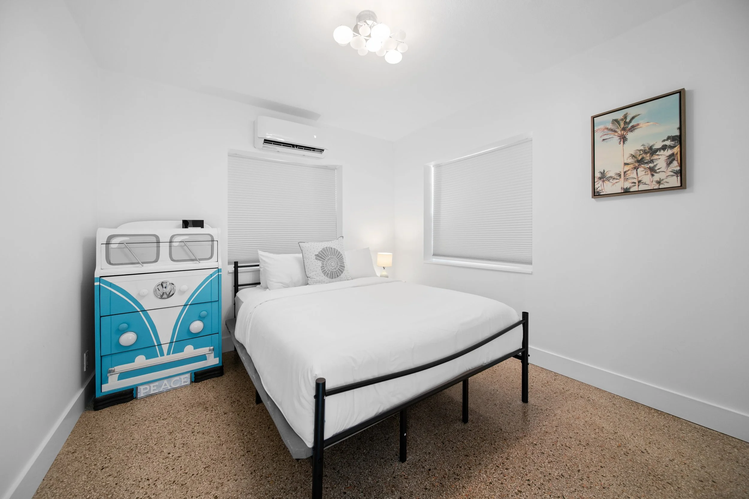 A bedroom with white walls, a bed with white sheets and pillows, a small lamp on a nightstand, and a palm tree picture on the wall. There is a window with blinds and a blue and white decorative item resembling a van with the words "PEACE" on it.