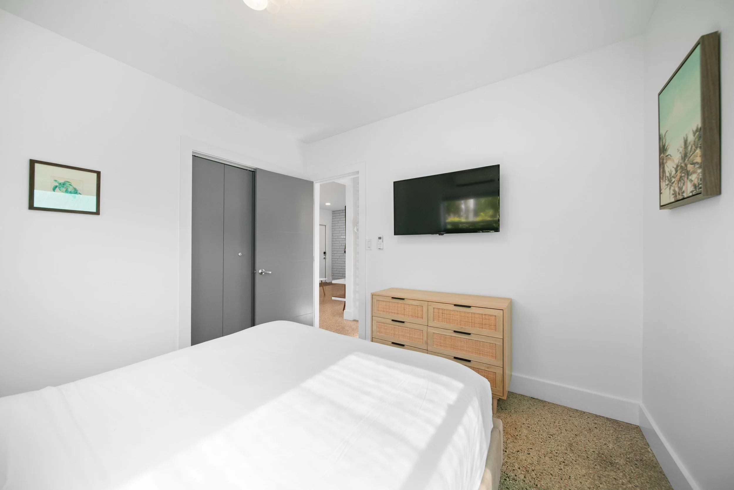 A bedroom with white walls, a bed with white bedding in the foreground, a wooden dresser, a wall-mounted flat-screen TV, and framed artwork, with an open door leading to a hallway.