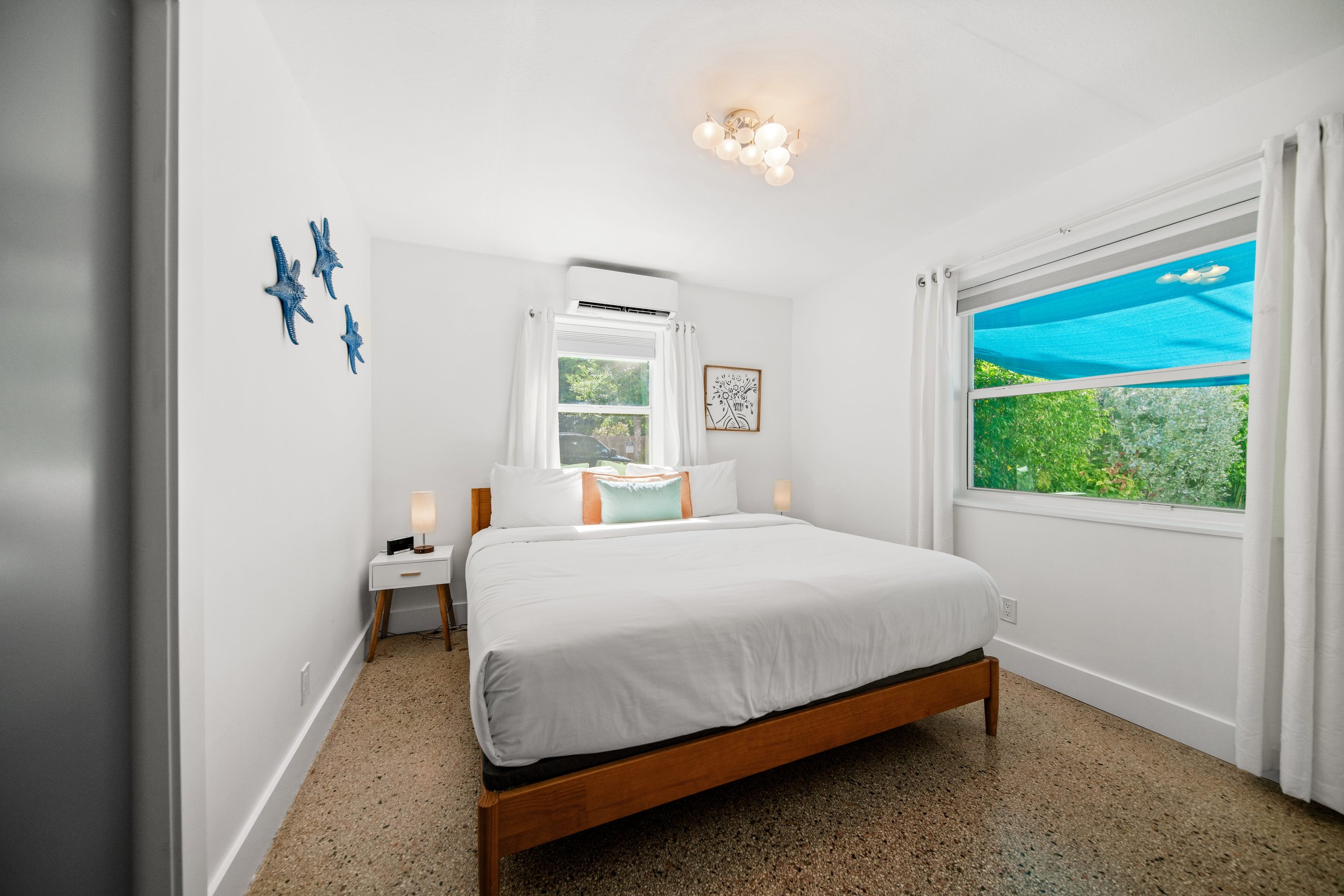 A bedroom with a wooden bed frame, white bedding, and multiple pillows. There are two small nightstands with lamps on either side of the bed. The room has white walls, a window with white curtains, and a large window or sliding door with an outdoor view of green trees and a blue awning. A decorative wall piece with blue starfish is visible on the left wall, and a framed art piece is on the far wall. A ceiling light fixture with multiple round bulbs is also visible.