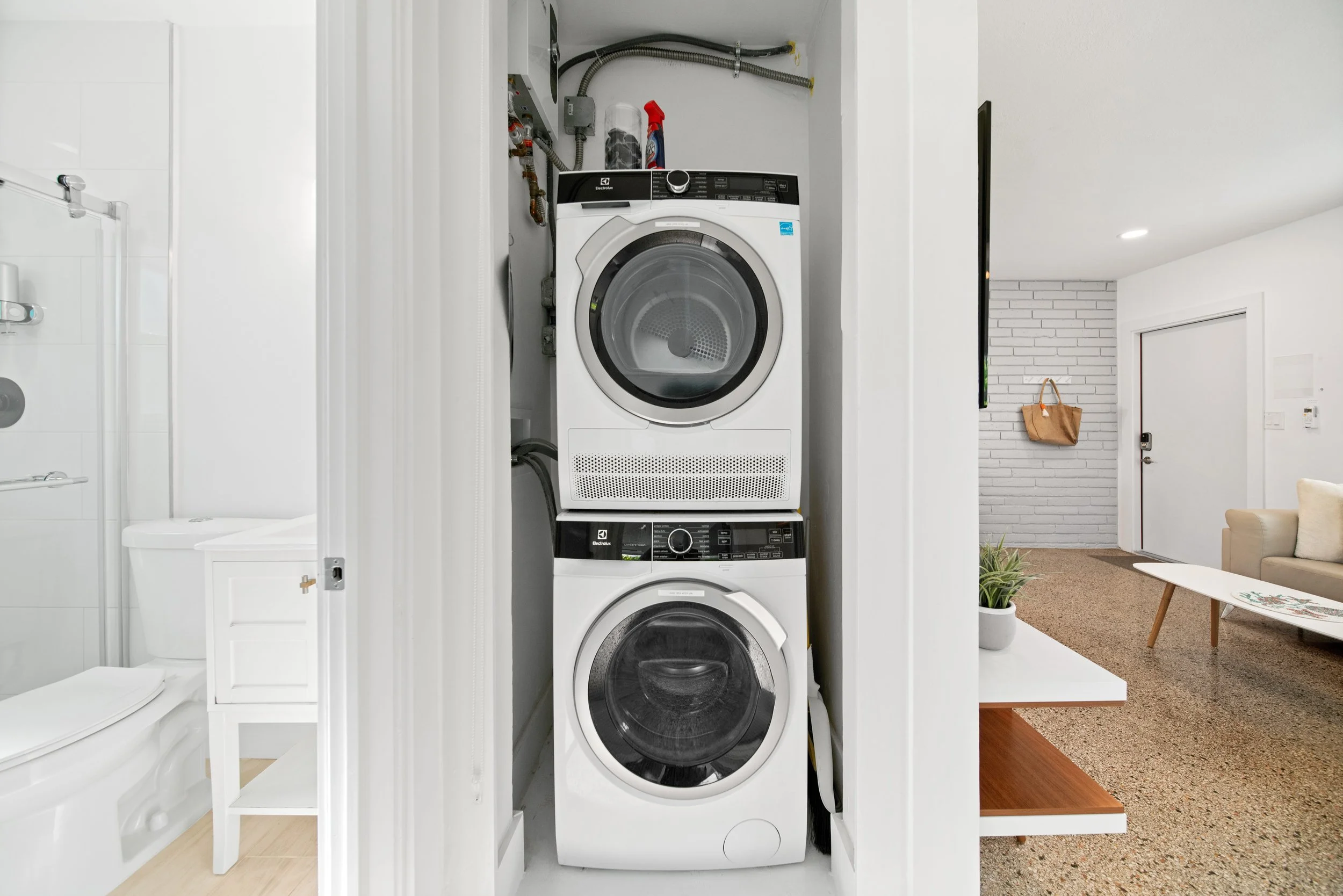 Stacked washer and dryer in a laundry closet with utility connections above, with part of a bathroom with white tiles and a shower on the left, and a living room with a brick accent wall, a door, a beige sofa, a small coffee table, and a potted plant
