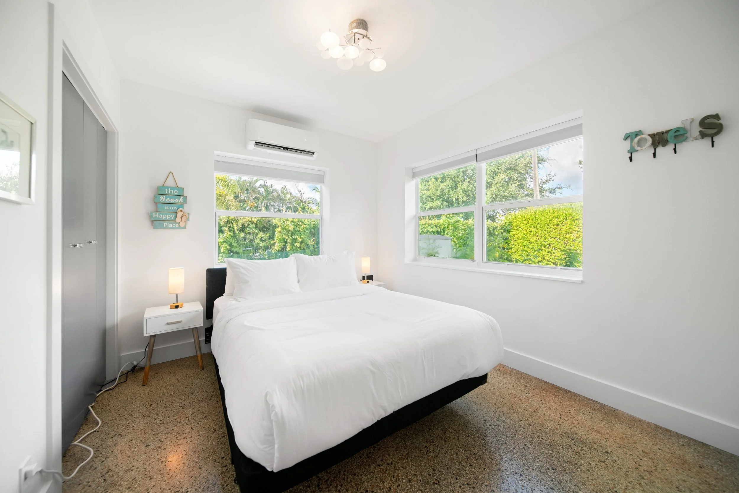 A bedroom with a large bed covered in white bedding, two bedside tables with lamps, and windows showing greenery outside. The walls are white, and there are decorative signs on the wall.