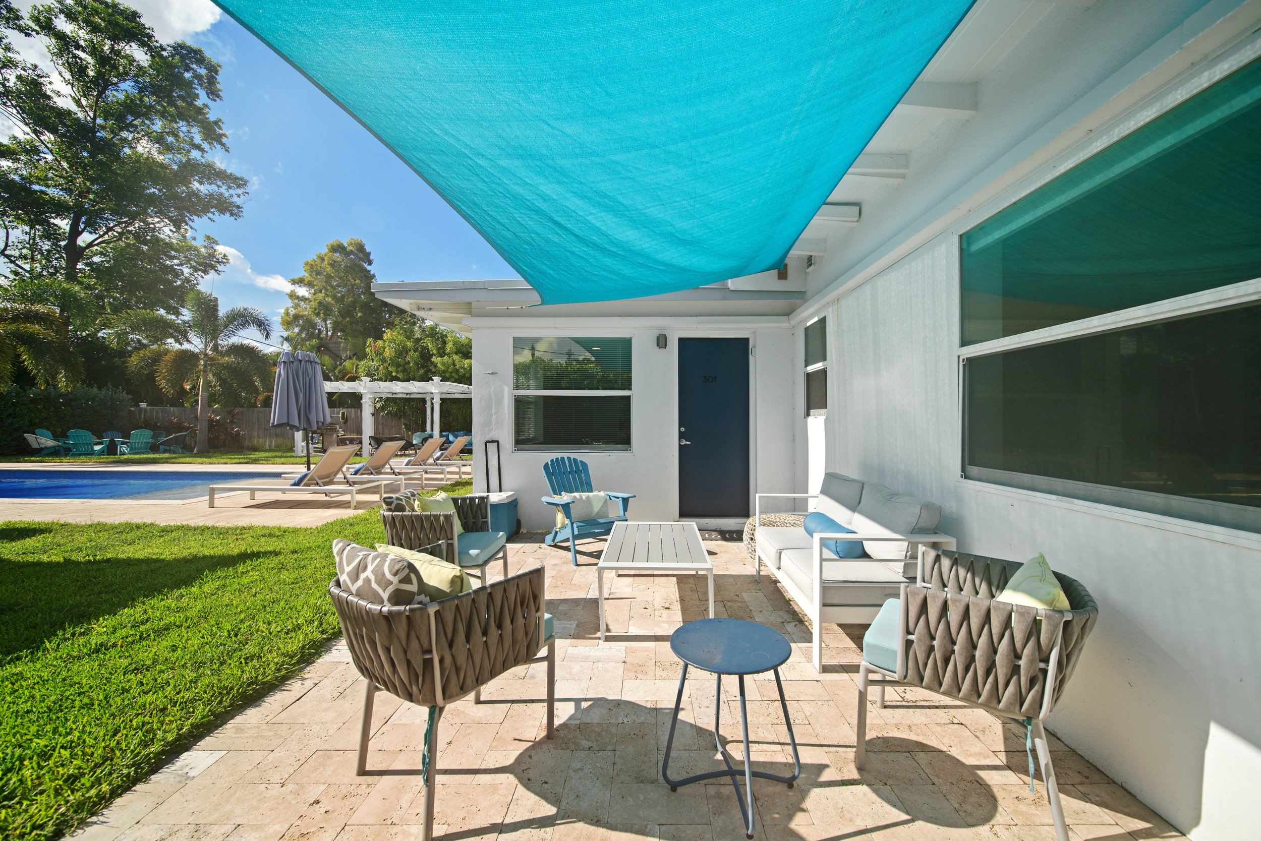 Exterior patio area with white furniture, blue cushions, and a blue shade sail overhead, adjacent to a house with large windows and a door, with a backyard pool, lawn, and trees in the background.
