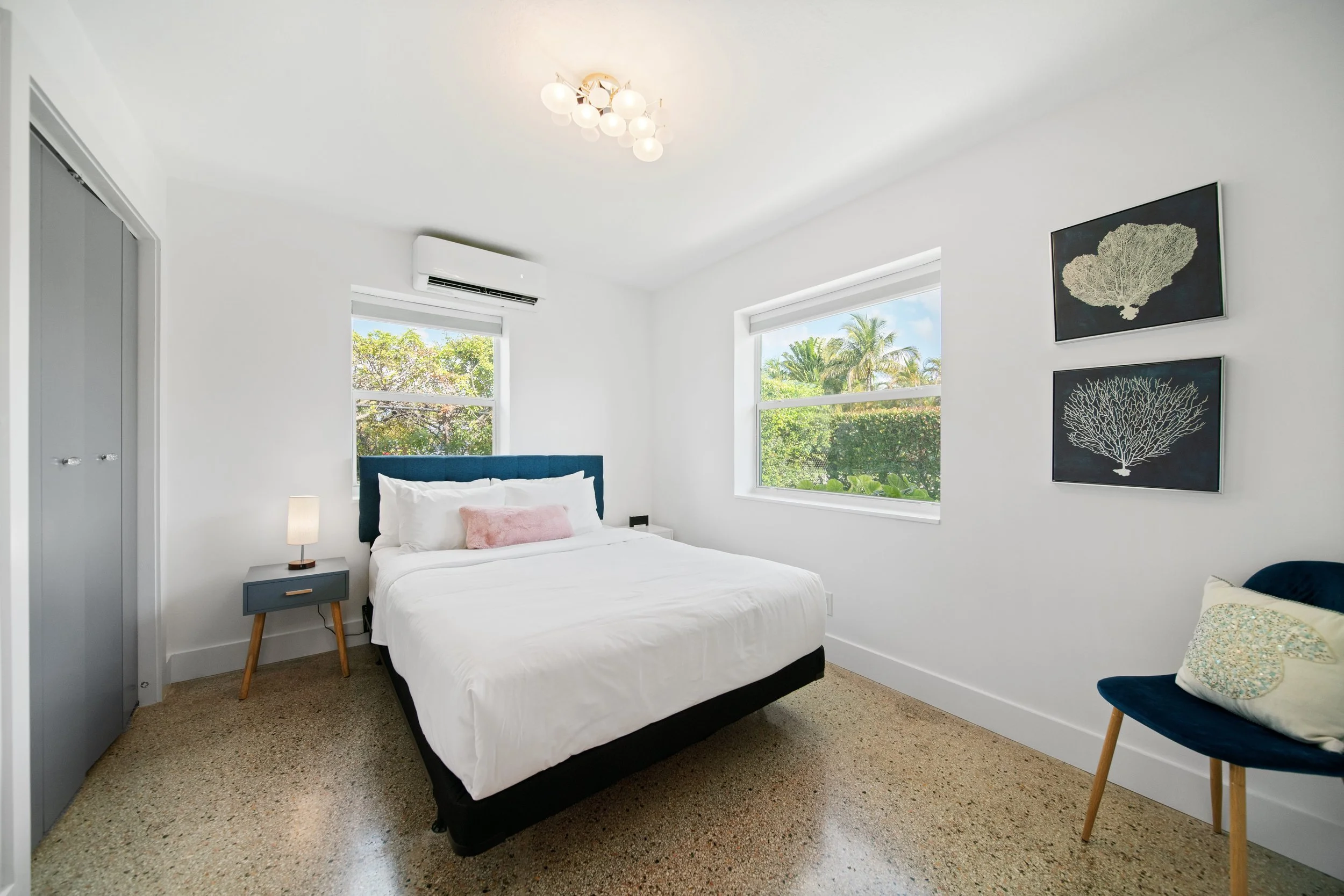 A bedroom with a bed, a small bedside table with a lamp, two windows showing greenery outside, and wall art depicting coral images.
