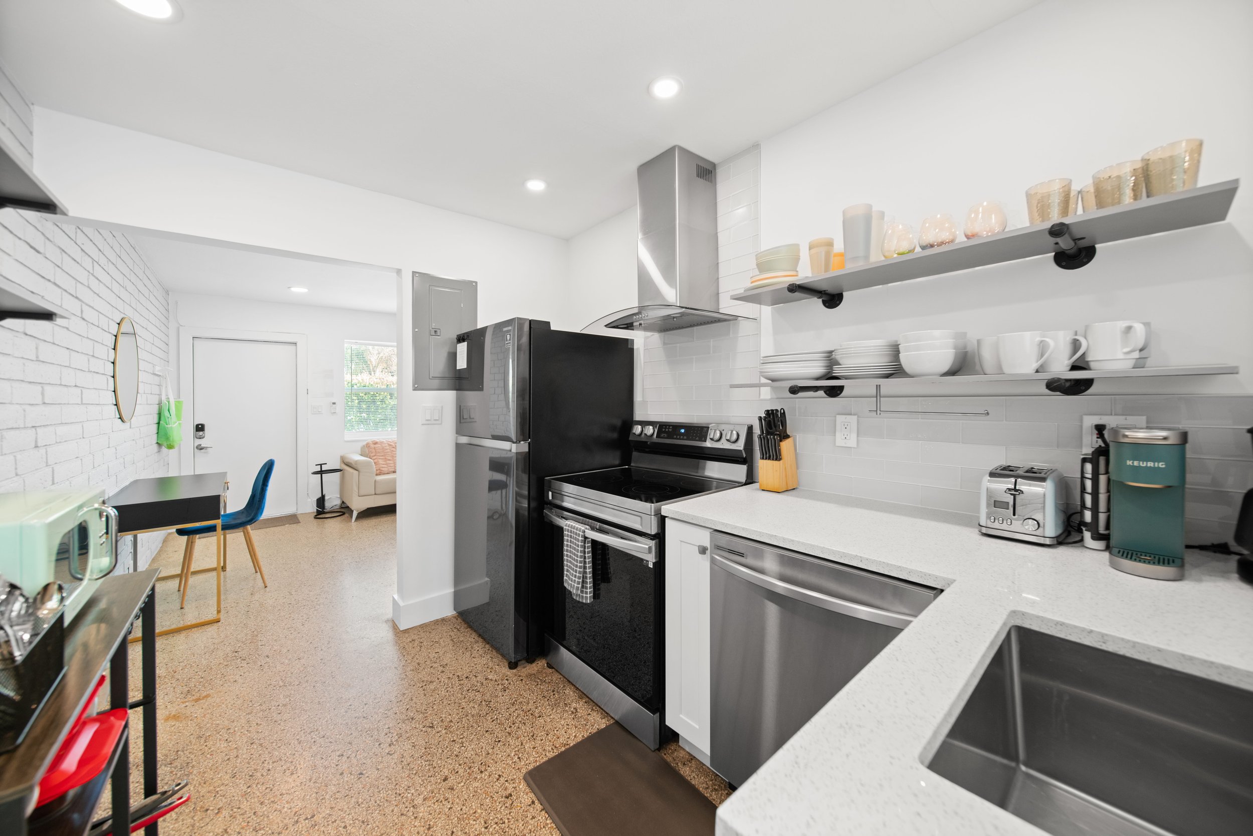 Modern kitchen with white cabinets, open shelving with bowls, glasses, and cups, black appliances, and a small dining area with a table and chairs.
