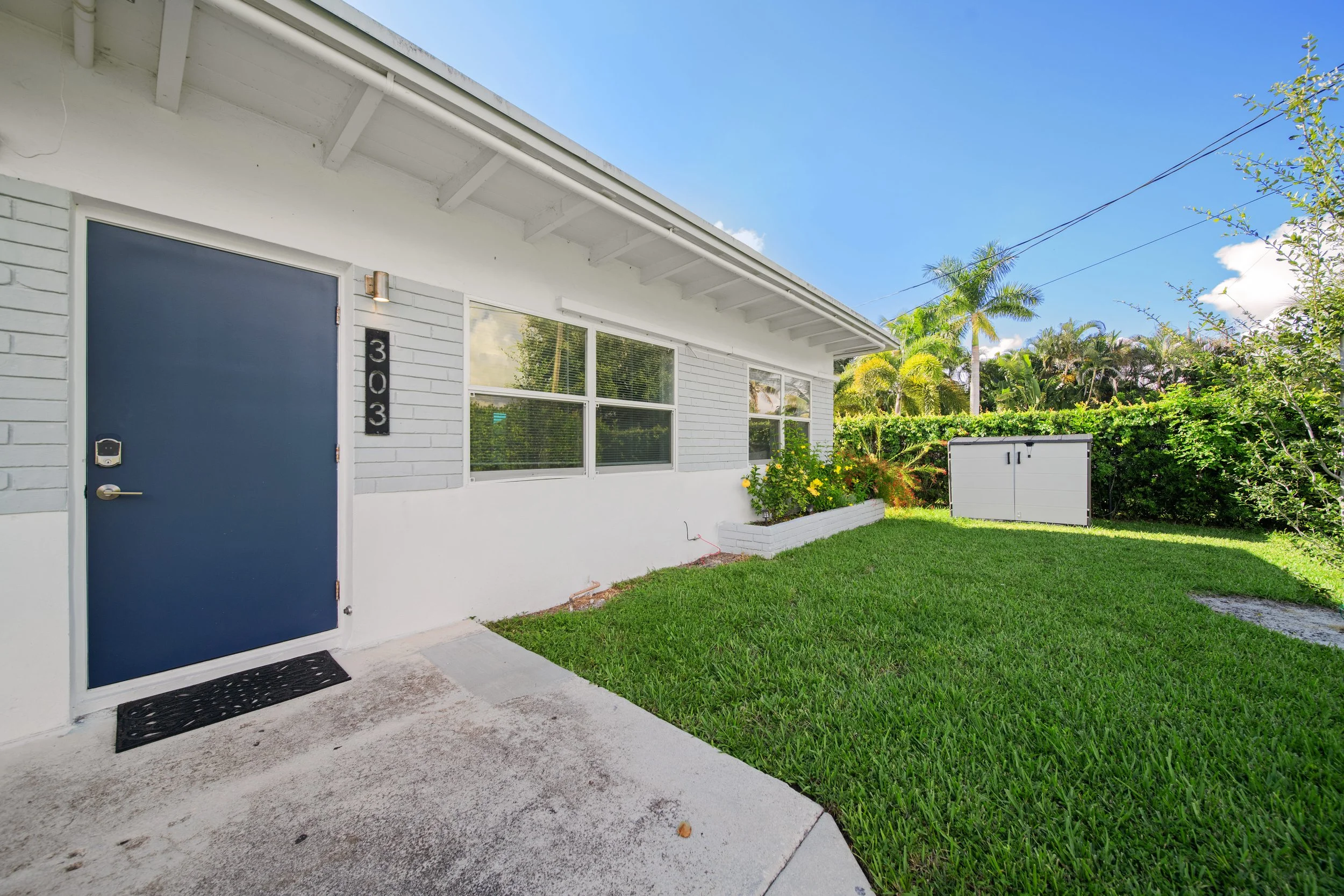 Front exterior of a modern house with a blue door, white brick walls, and large windows, surrounded by a well-maintained lawn, lush greenery, and palm trees, under a bright blue sky.