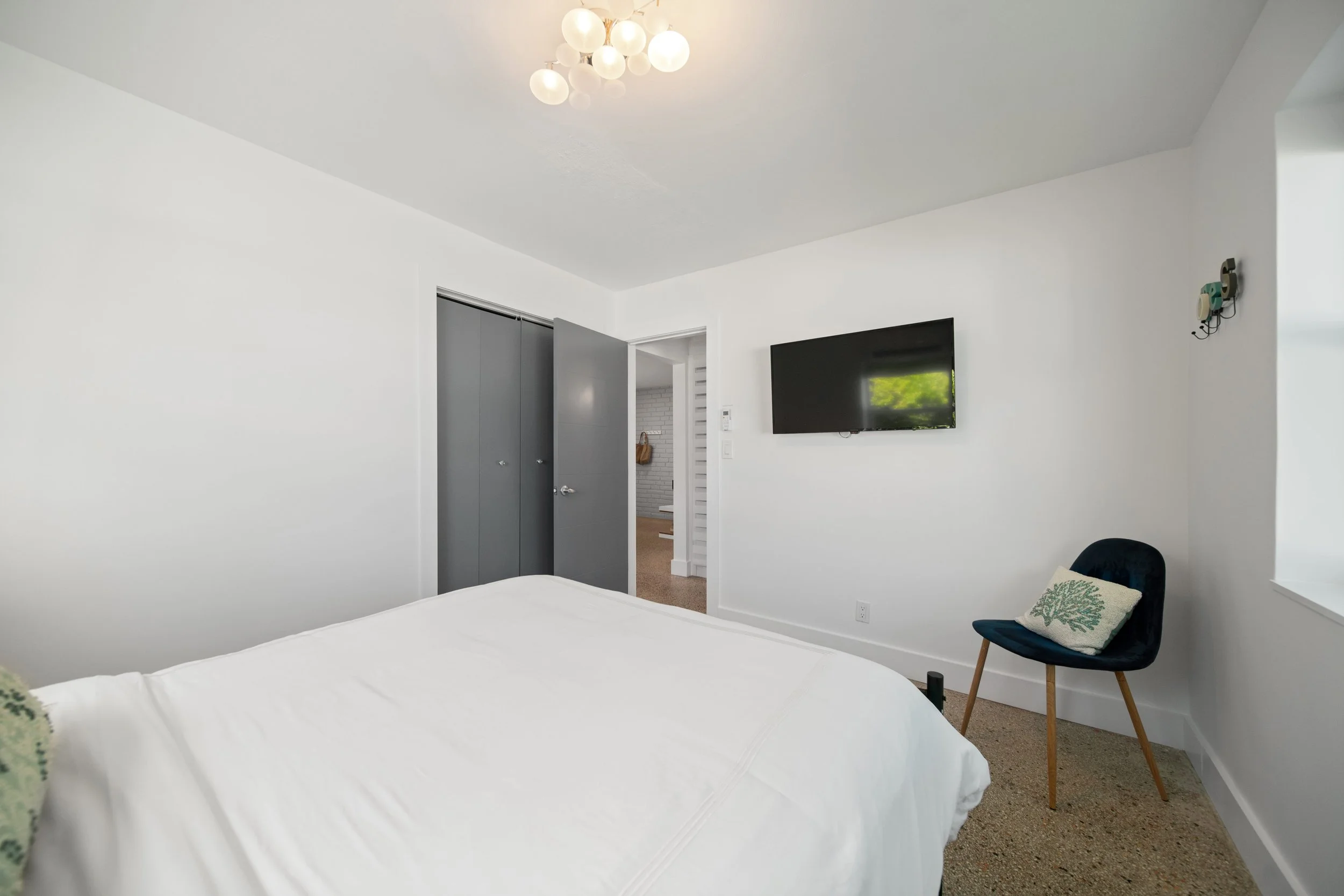 A bedroom with white walls and ceiling, ceiling light fixture with multiple white globes, wall-mounted TV, a blue chair with a decorative pillow, and an open gray door leading to a hallway.