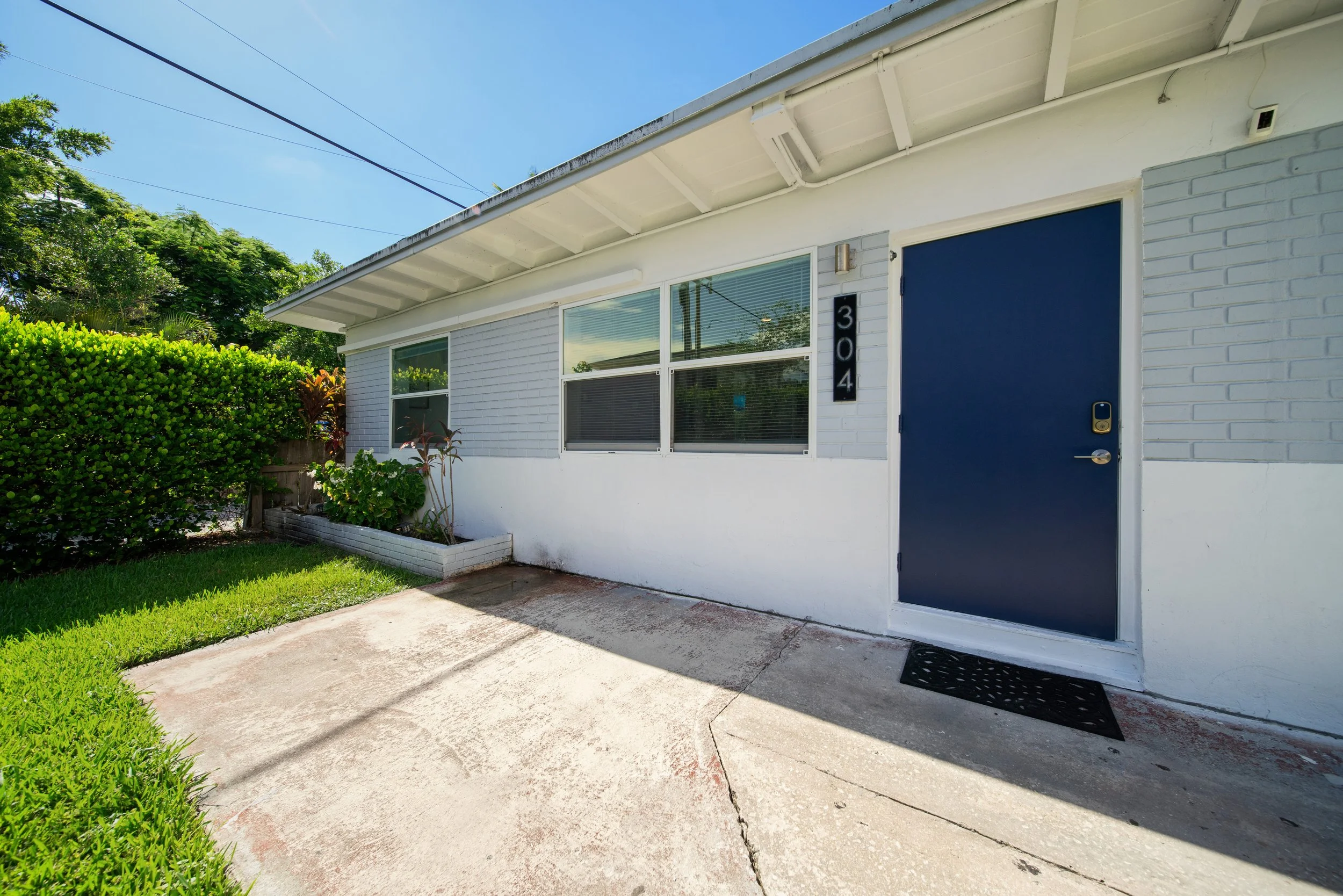 Front of a white house with a blue door, a small garden area, and a concrete walkway, under a clear blue sky.