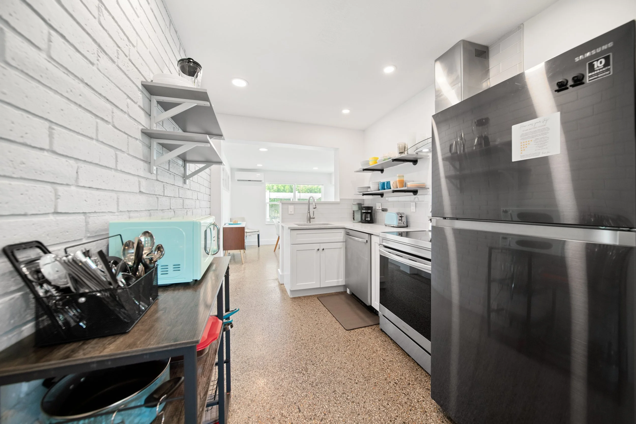 A modern kitchen with white cabinets, stainless steel appliances, open shelving with dishes, and a white brick wall with floating shelves.
