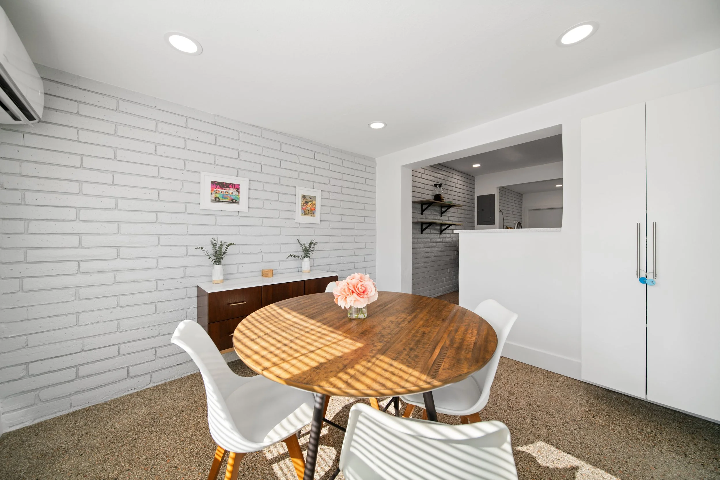 Modern dining area with a round wooden table, white chairs, a pink flower centerpiece, white brick wall with artwork, and a kitchen pass-through with shelves.