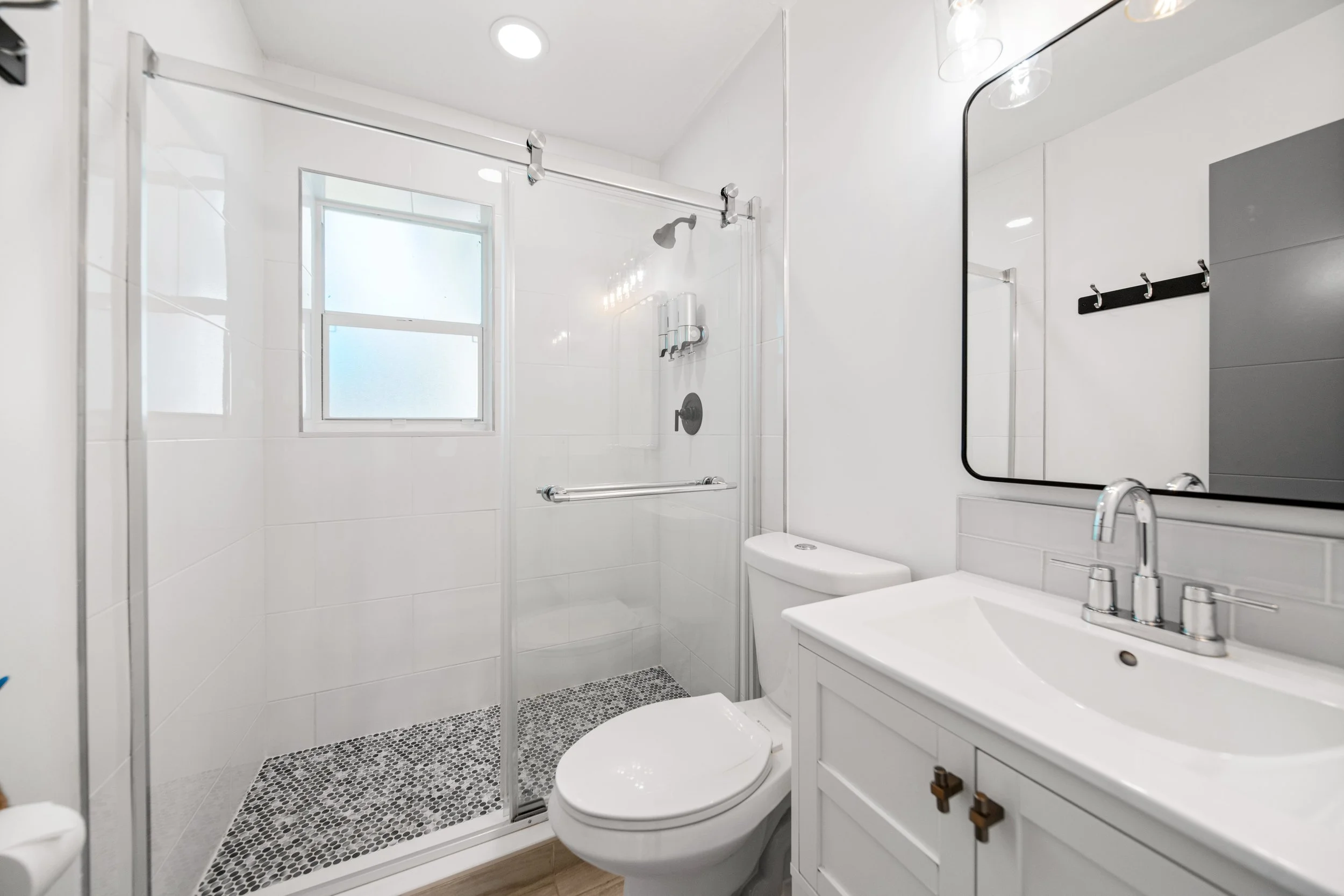 A modern bathroom with white walls, a glass shower enclosure, and a window. It features a white toilet, a white vanity with a sink, and a large mirror. The shower has a rain showerhead, and there are small black and white mosaic tiles on the shower f