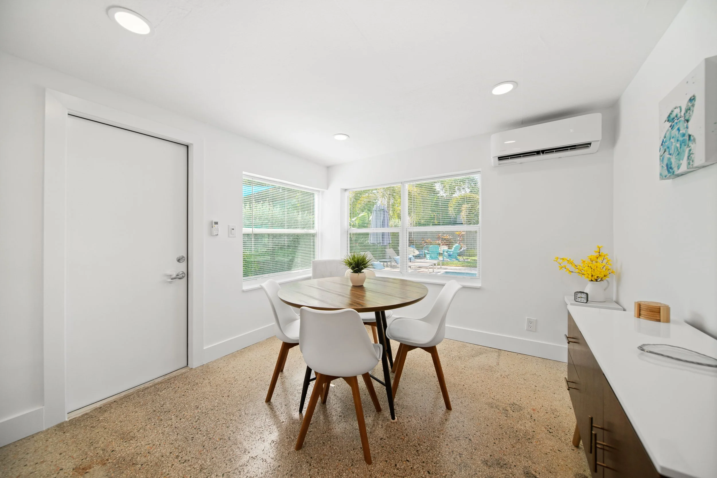 Dining area with round wooden table, four white chairs, large windows with blinds, air conditioner, some yellow flowers on a side table, and abstract artwork on the wall.