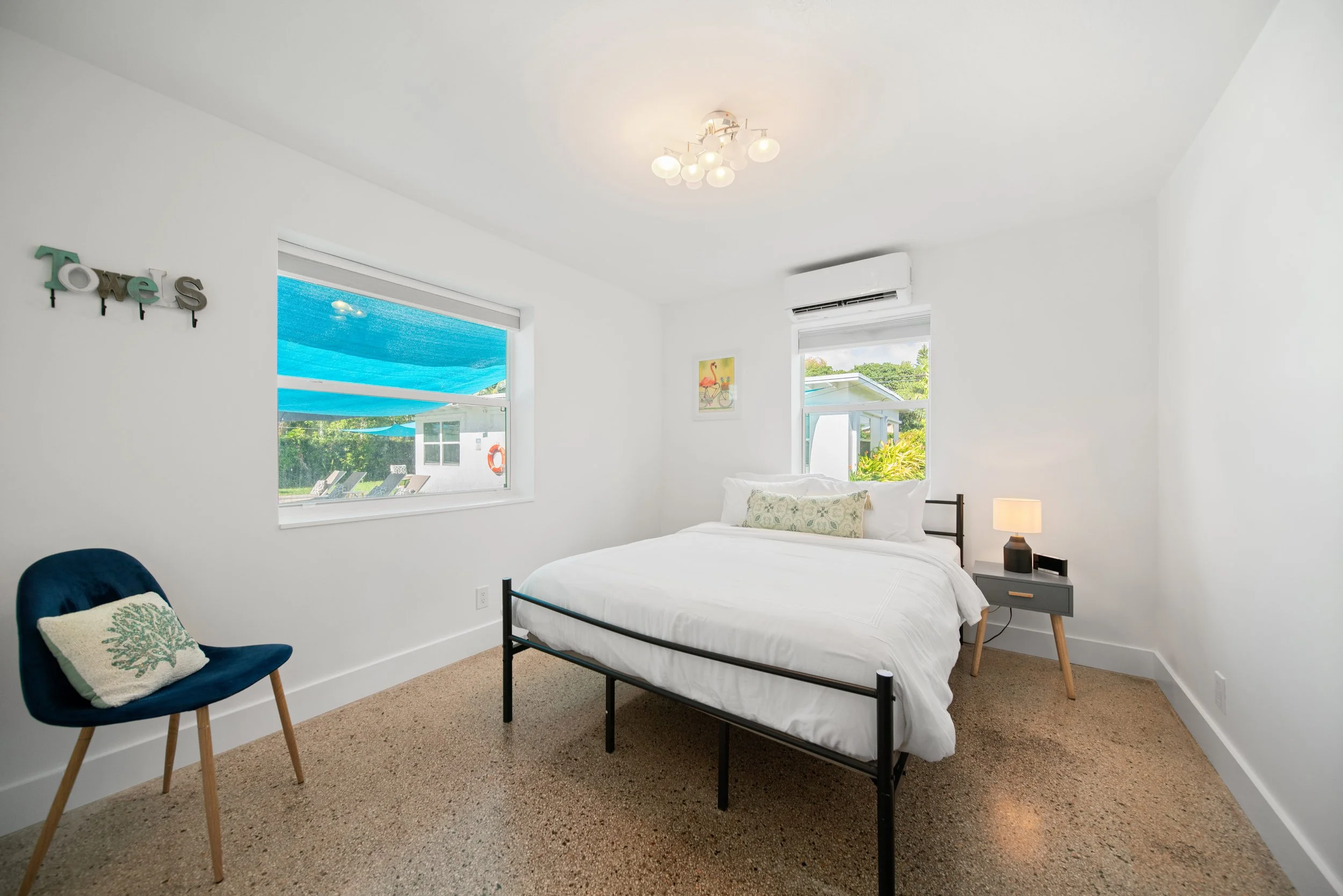A bedroom with white walls, a black metal bed, two windows with views of outdoor greenery, a modern ceiling light fixture, a navy blue chair with a decorative pillow, a gray nightstand with a lamp, and wall decor including a "Towels" sign and a color