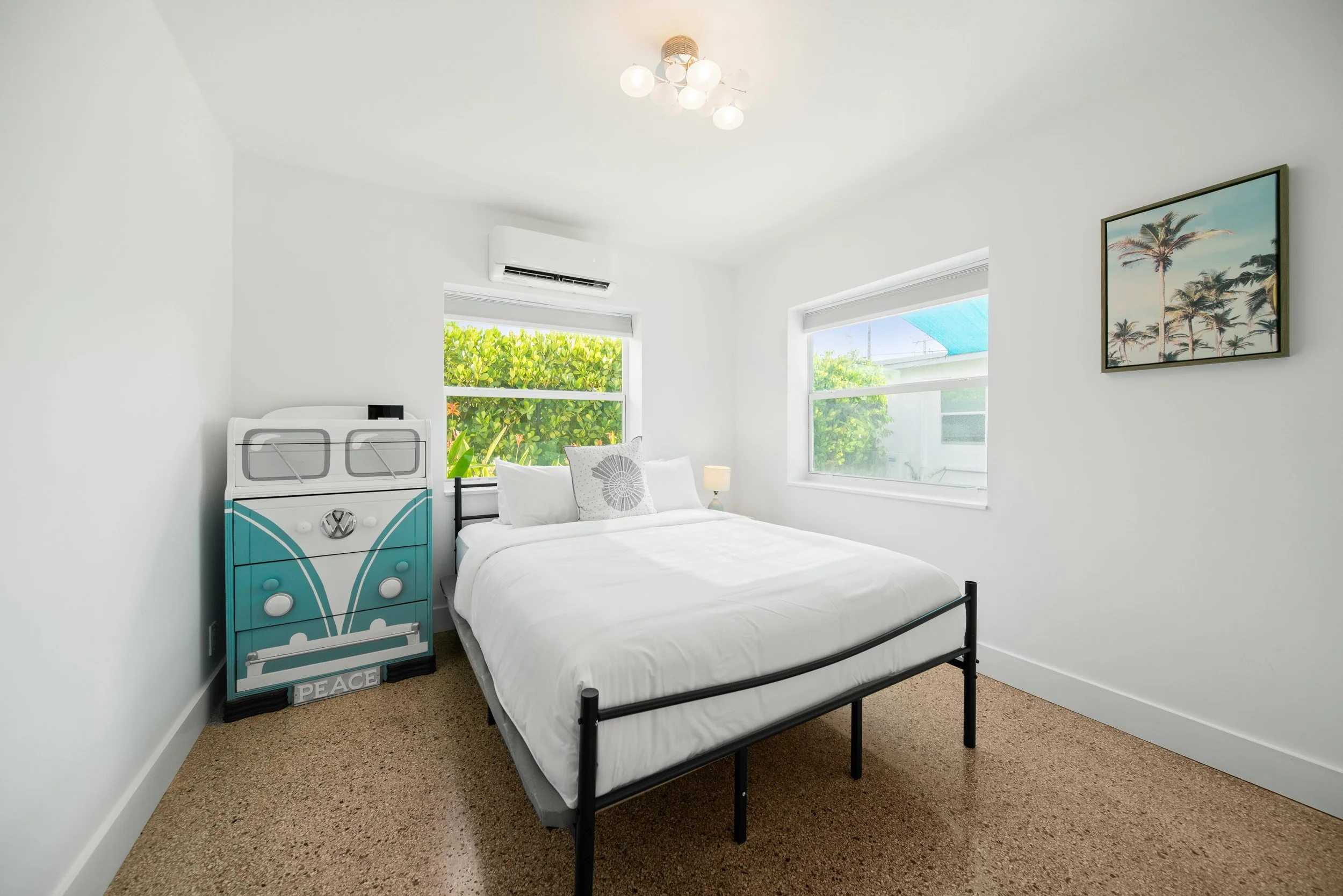 A bedroom with white walls and a bed with white bedding. Two windows with green foliage outside, a framed picture of palm trees, and a ceiling light fixture with round bulbs. There is a blue and white camper van-shaped nightstand with the words "PEAC