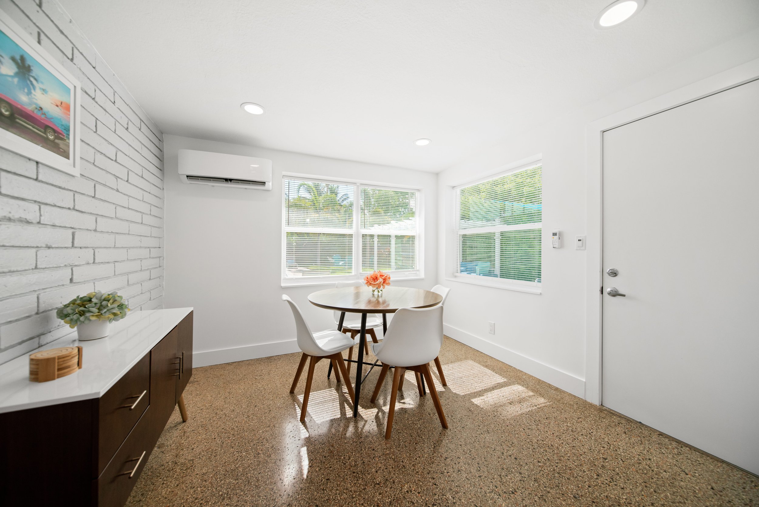 A small dining area with a round wooden table and four white chairs, large windows with blinds, a white brick wall with a colorful painting, a sideboard with plants, and an air conditioning unit, in a brightly lit room.