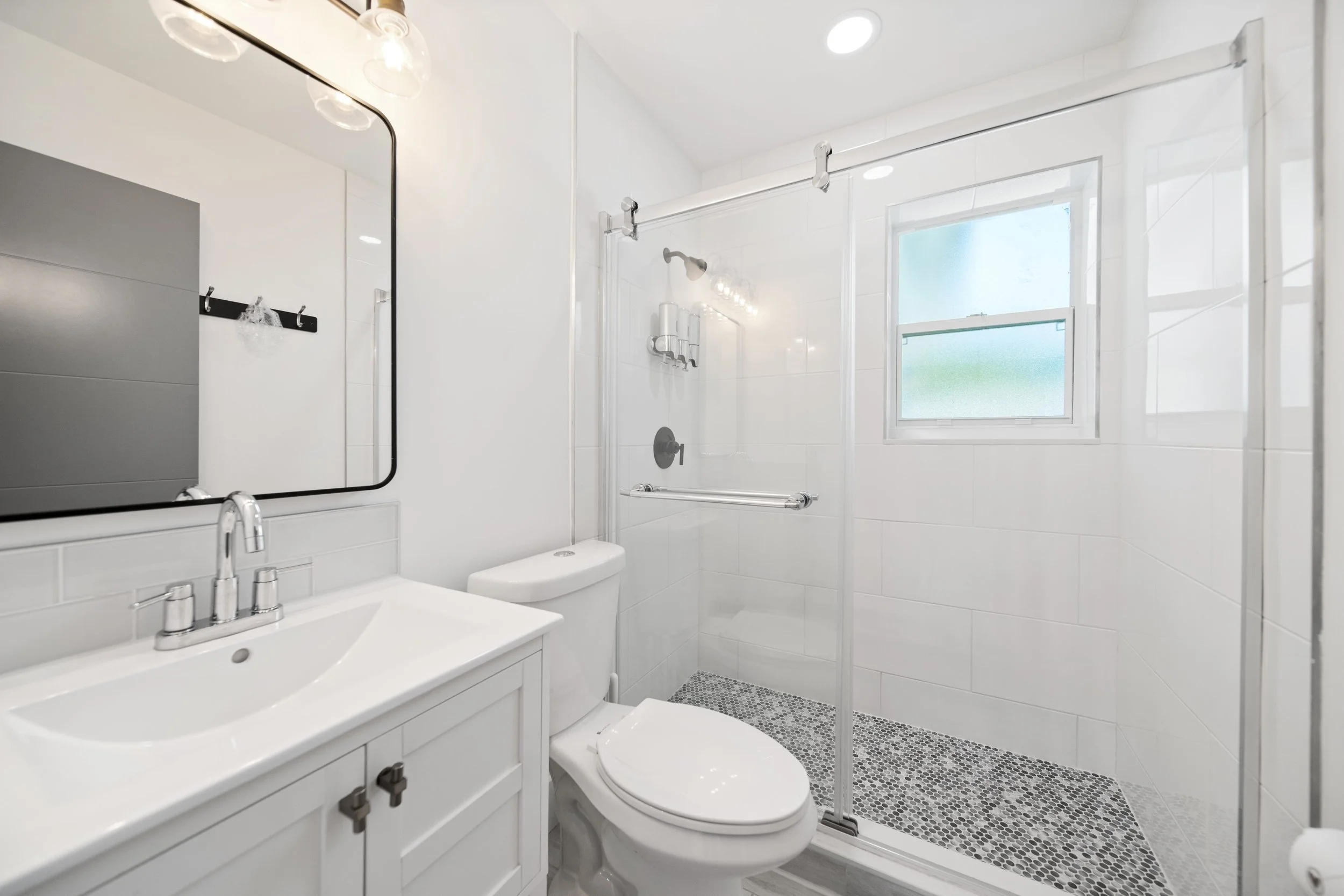 A modern bathroom with a white vanity, mirror, toilet, and a glass-enclosed shower. The bathroom has white tiled walls, a small frosted window, and a gray pebble tile floor in the shower area.