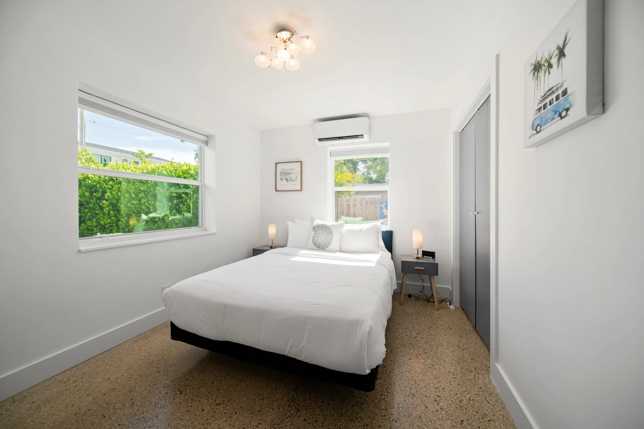 A bedroom with white walls, a bed with white linens, two small bedside tables with lamps, two windows letting in natural light, and minimal wall art including a framed picture and a canvas with palm trees and a vehicle, with carpeted floor.