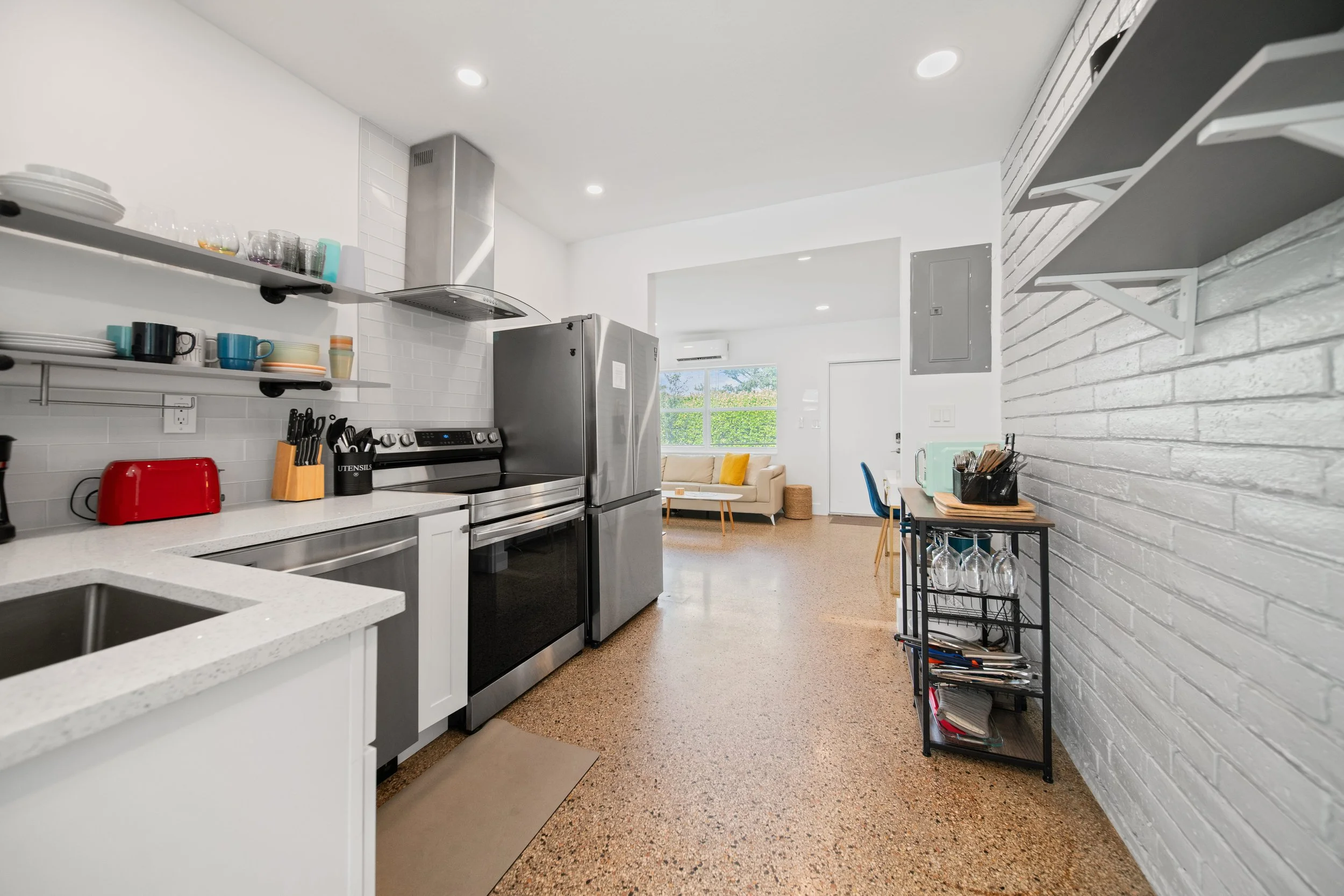 Modern kitchen with white countertops, stainless steel appliances, open shelves with dishes and glasses, and a sitting area with a sofa visible through an open doorway.