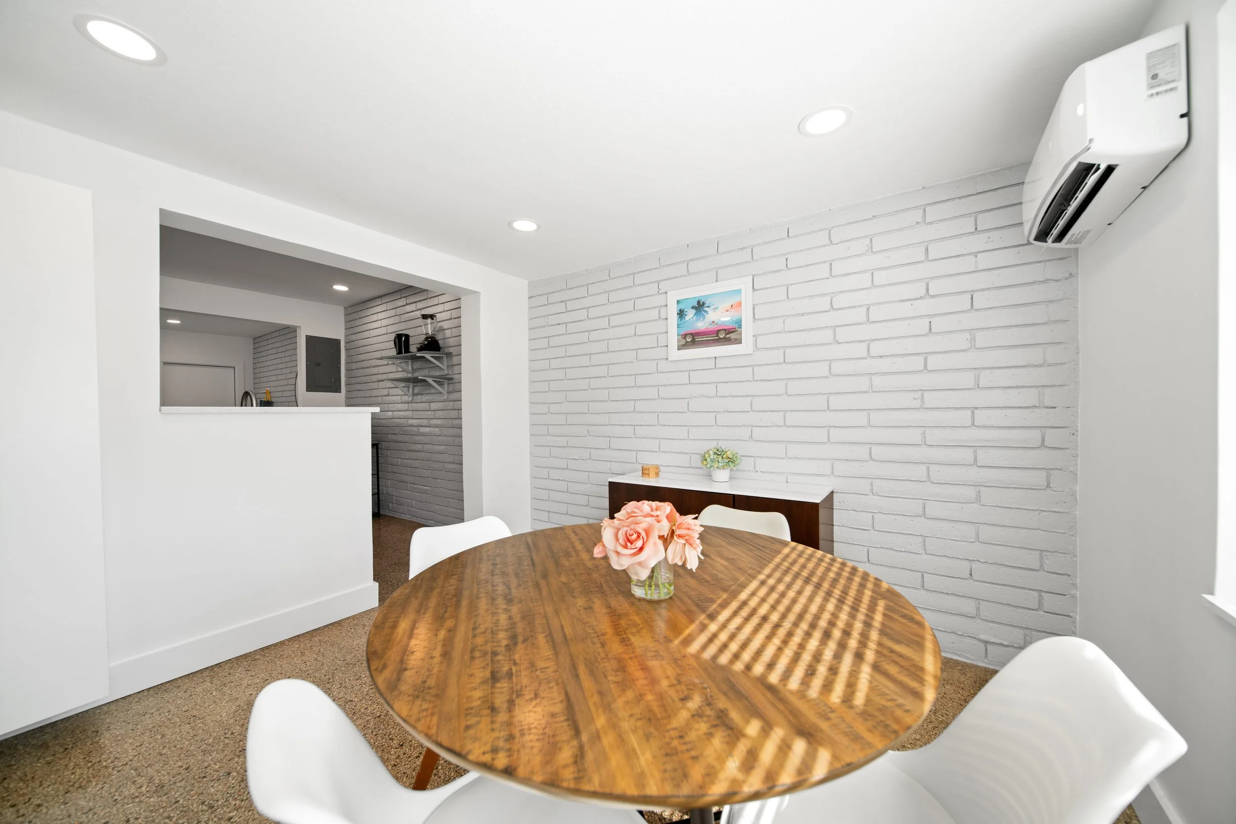 Dining room with a wooden table surrounded by white chairs, a vase with pink roses on the table, white brick walls, a framed colorful picture of a car and palm trees, and an air conditioning unit on the wall.