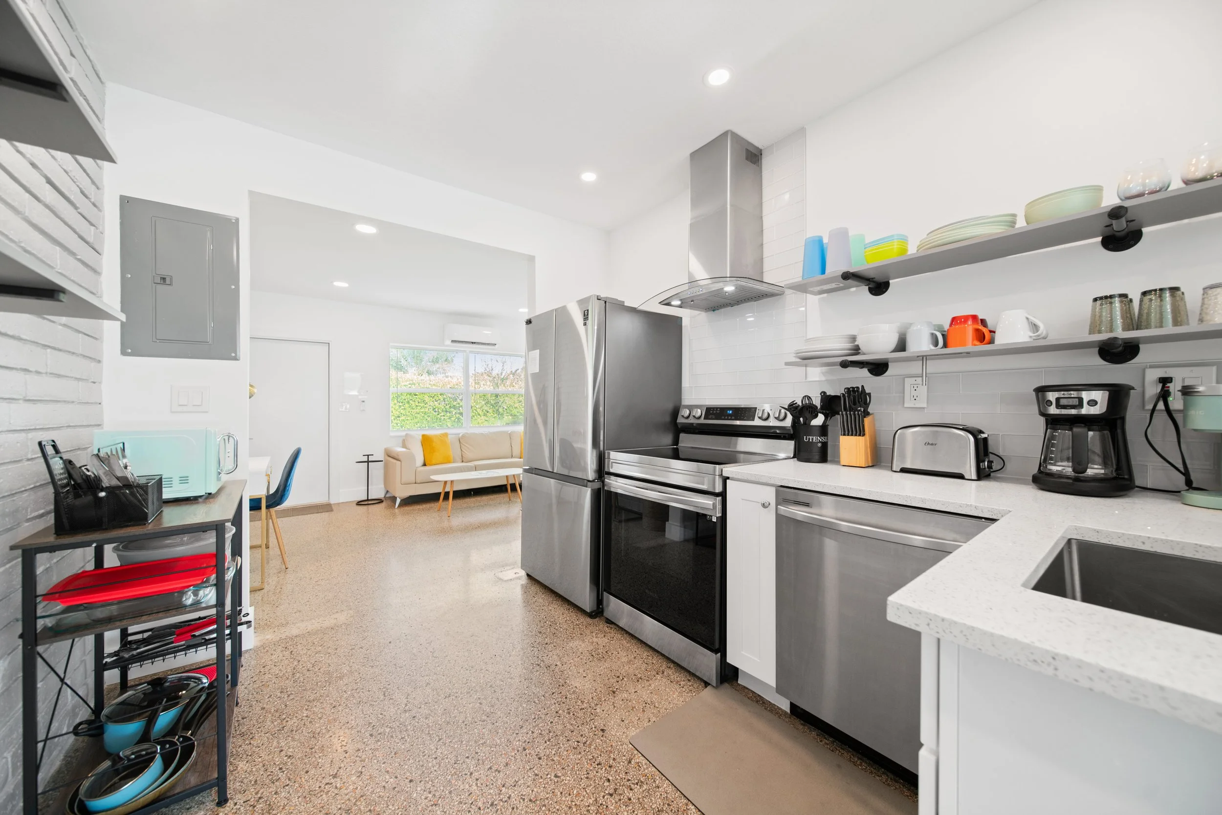 Modern kitchen with white cabinets, stainless steel appliances, open shelving with dishes and bowls, kitchen appliances on the counter, and a view into a living area with a beige sofa and a large window.