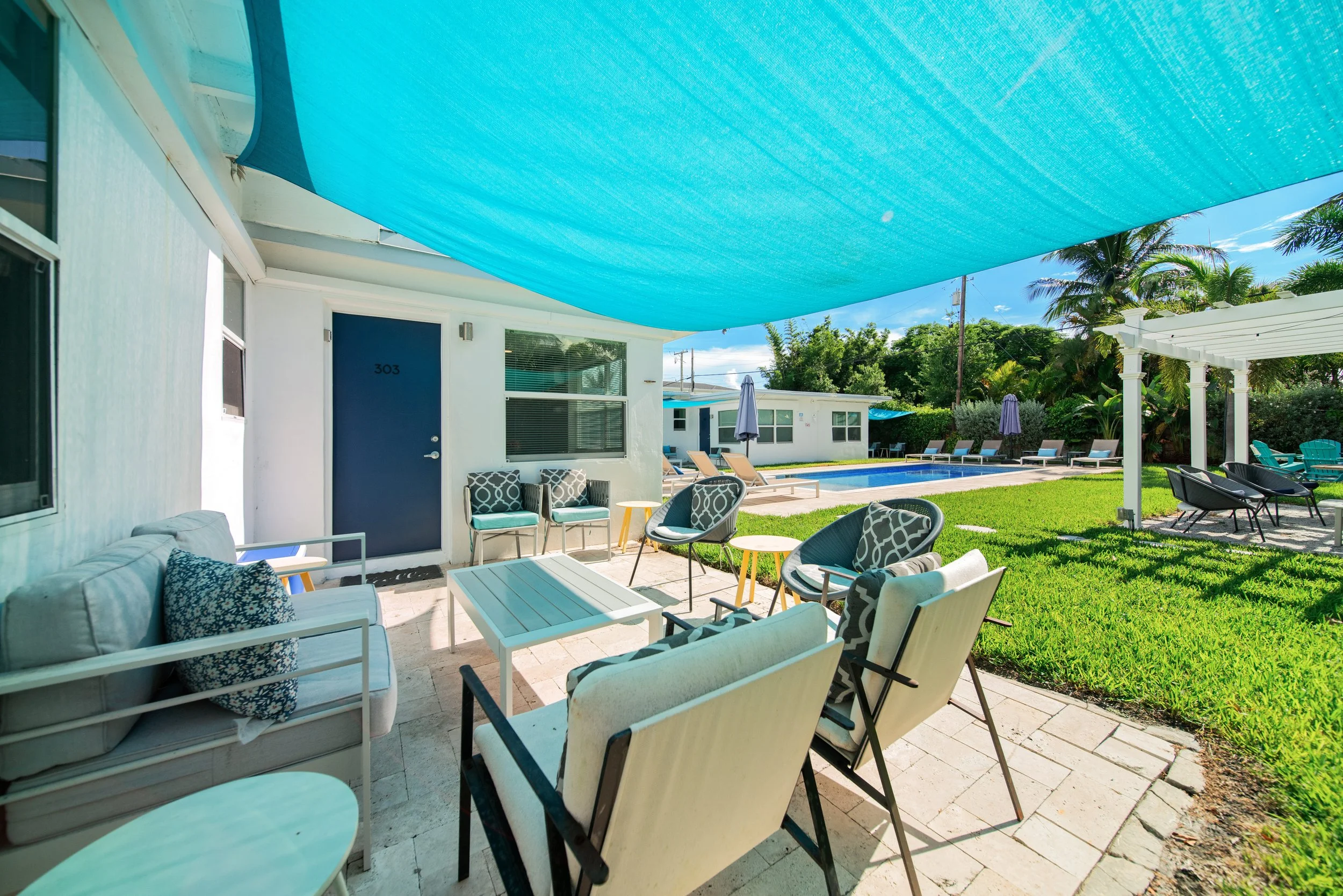 A backyard patio area with outdoor furniture, a swimming pool, and a large blue shade sail covering the space.